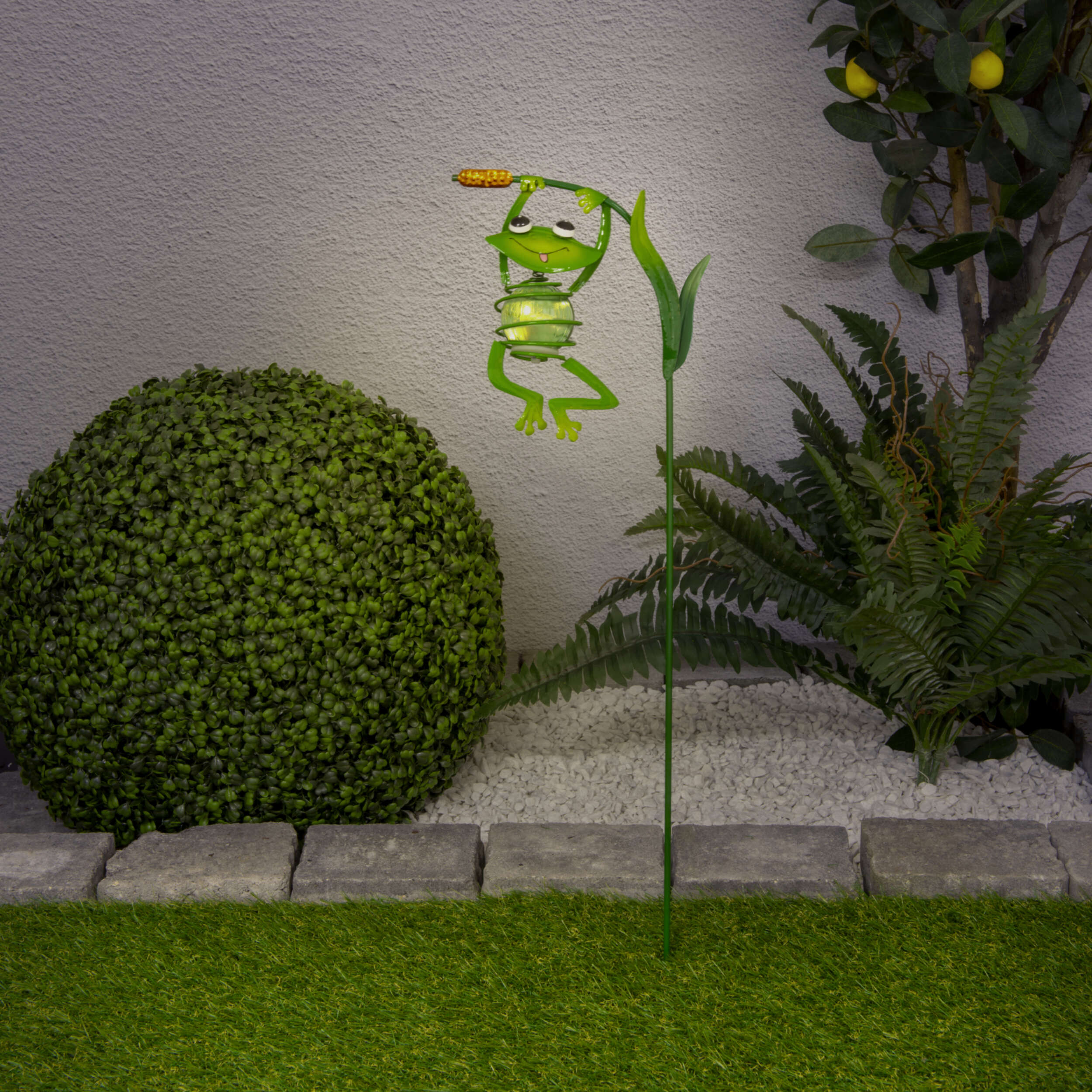 Ein grüner froschförmiger Gartenpfahl mit einem Licht in einem Garten mit Pflanzen.