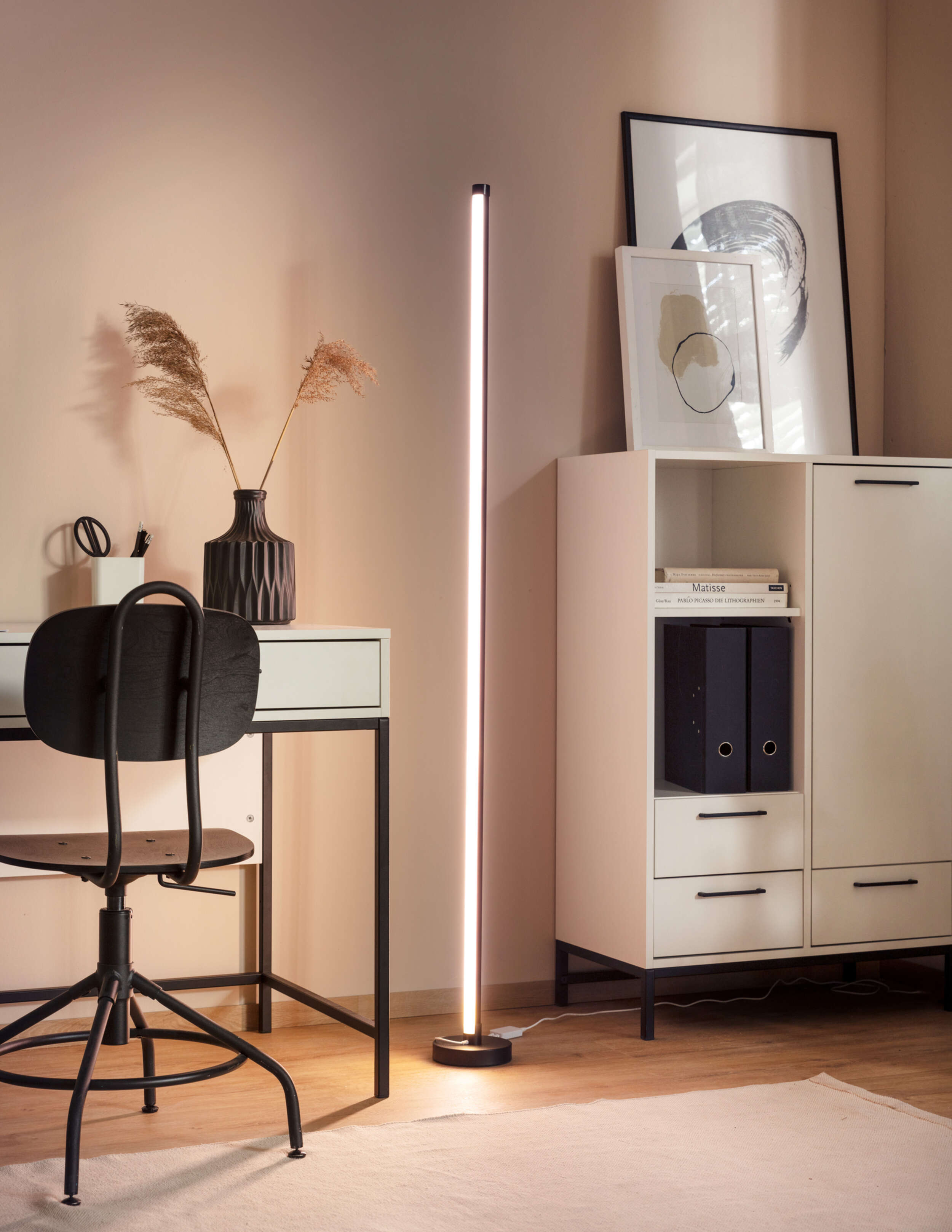 Eine hohe, beleuchtete, moderne Stehlampe in einer Raumsituation mit Schreibtisch und Stuhl.