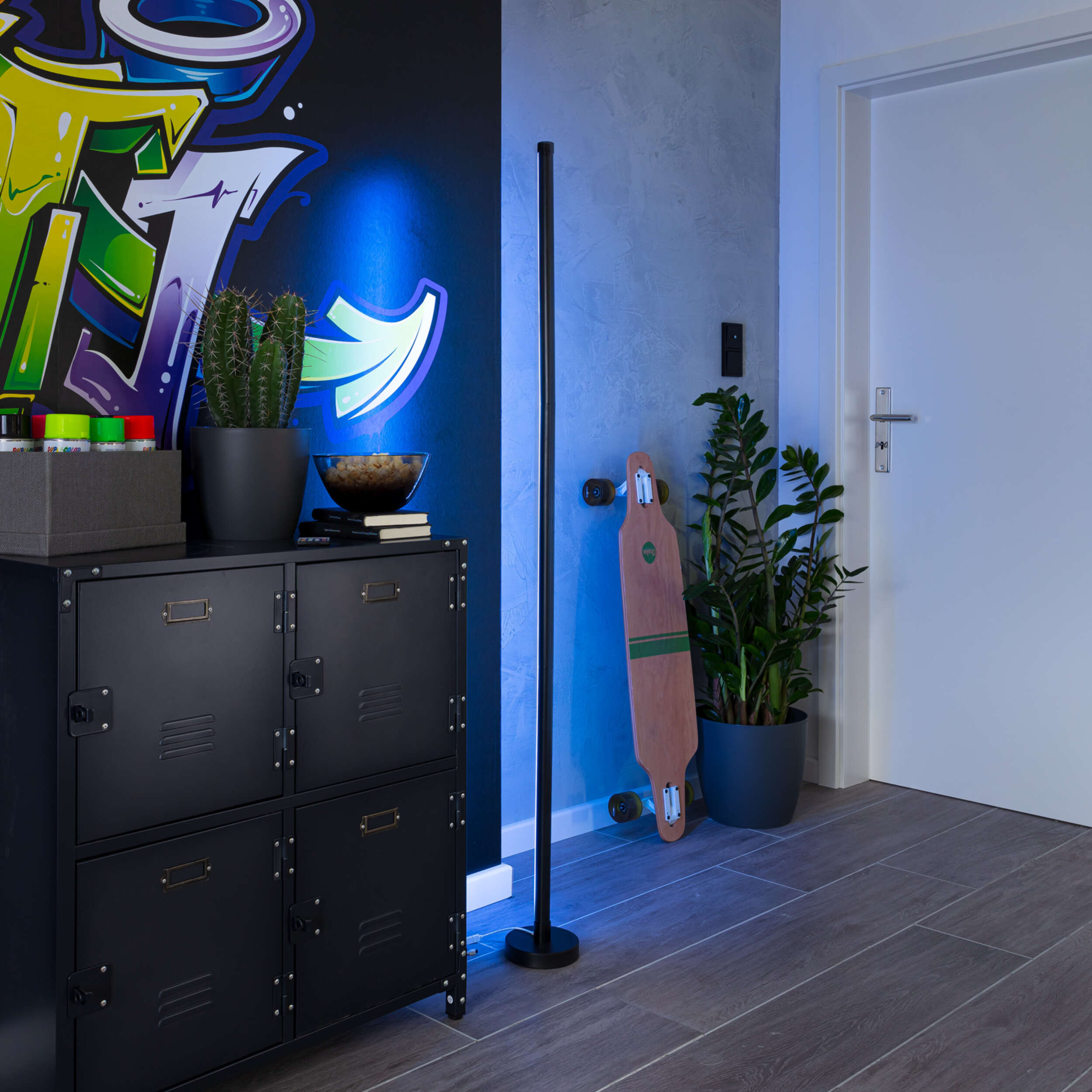 Innenraum mit schwarzem Schrank, Stehlampe, Graffiti-Kunst und Skateboard an einer Wand.