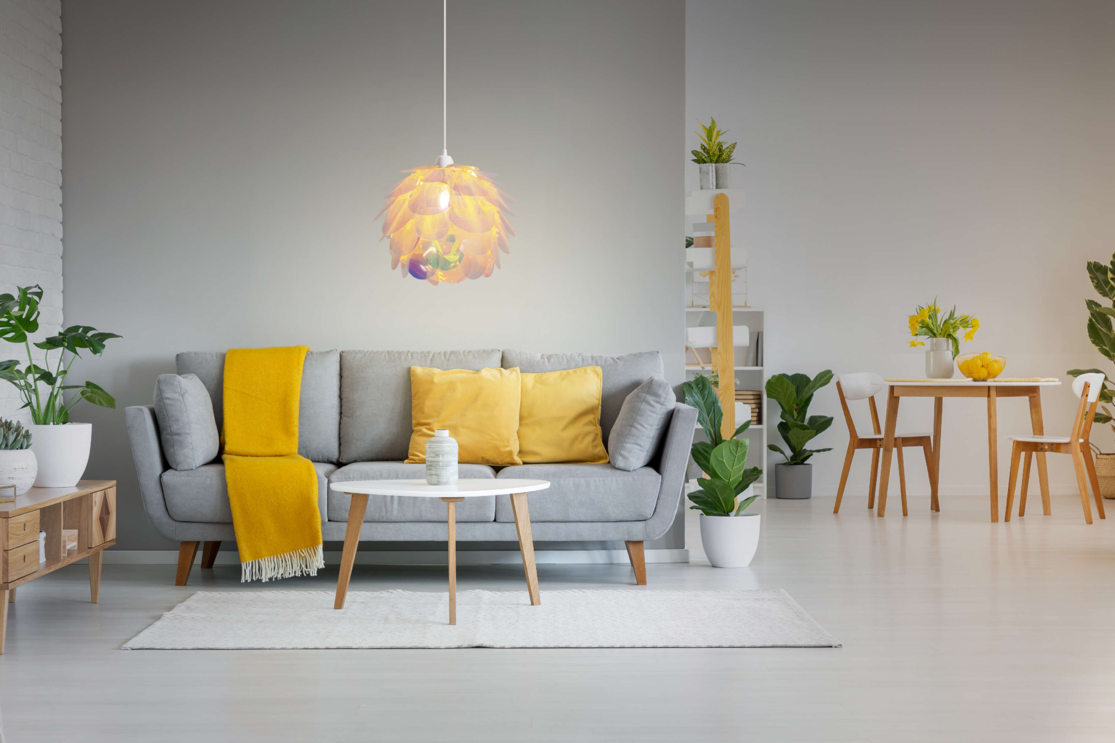 Wohnzimmer mit grauem Sofa, gelben Kissen und Decke, weißem Tisch und Pflanzen. Helle Beleuchtung.
