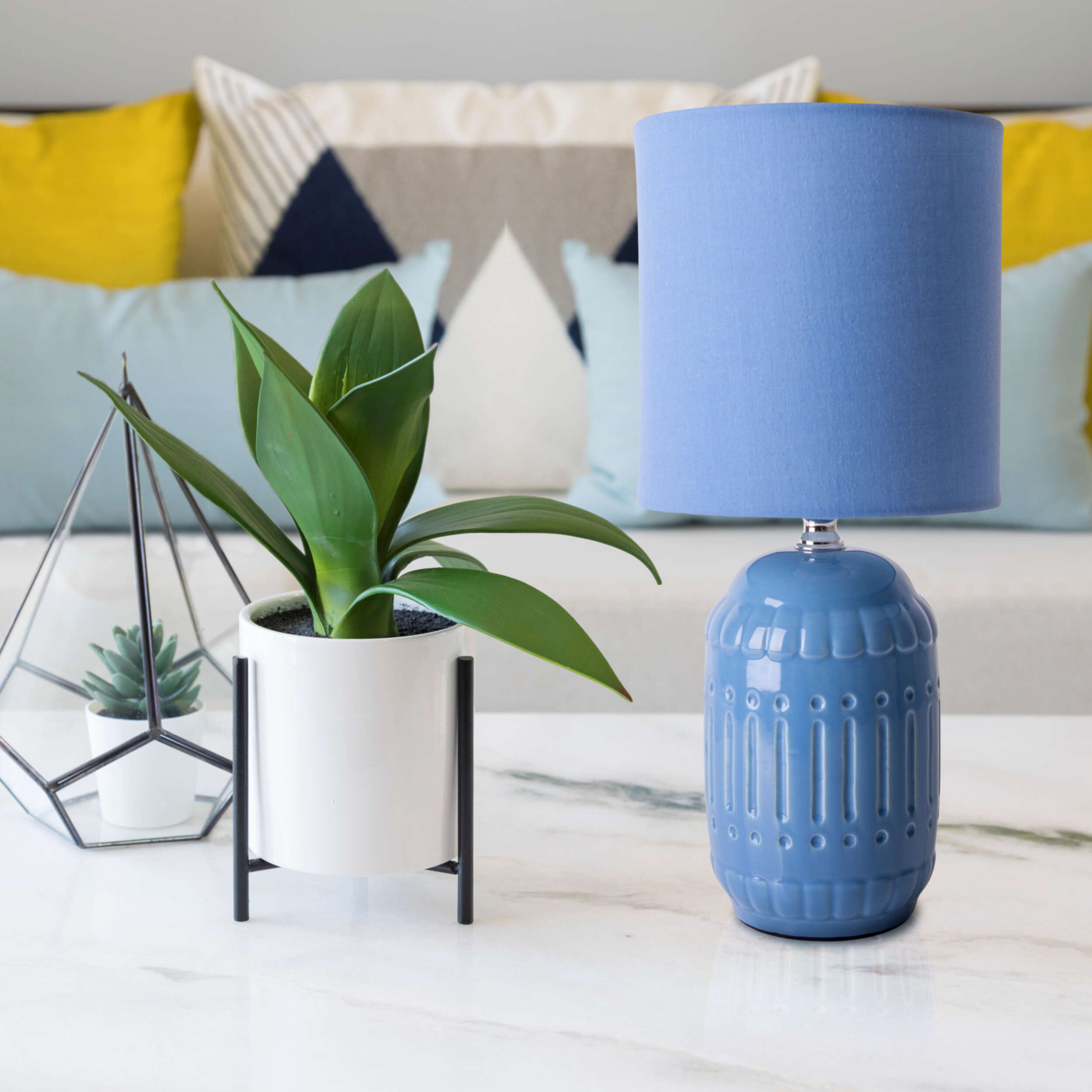 Blaue Lampe auf einem weißen Tisch. Eine Pflanze in einem weißen Topf und eine geometrische Dekoration stehen auf dem Tisch.