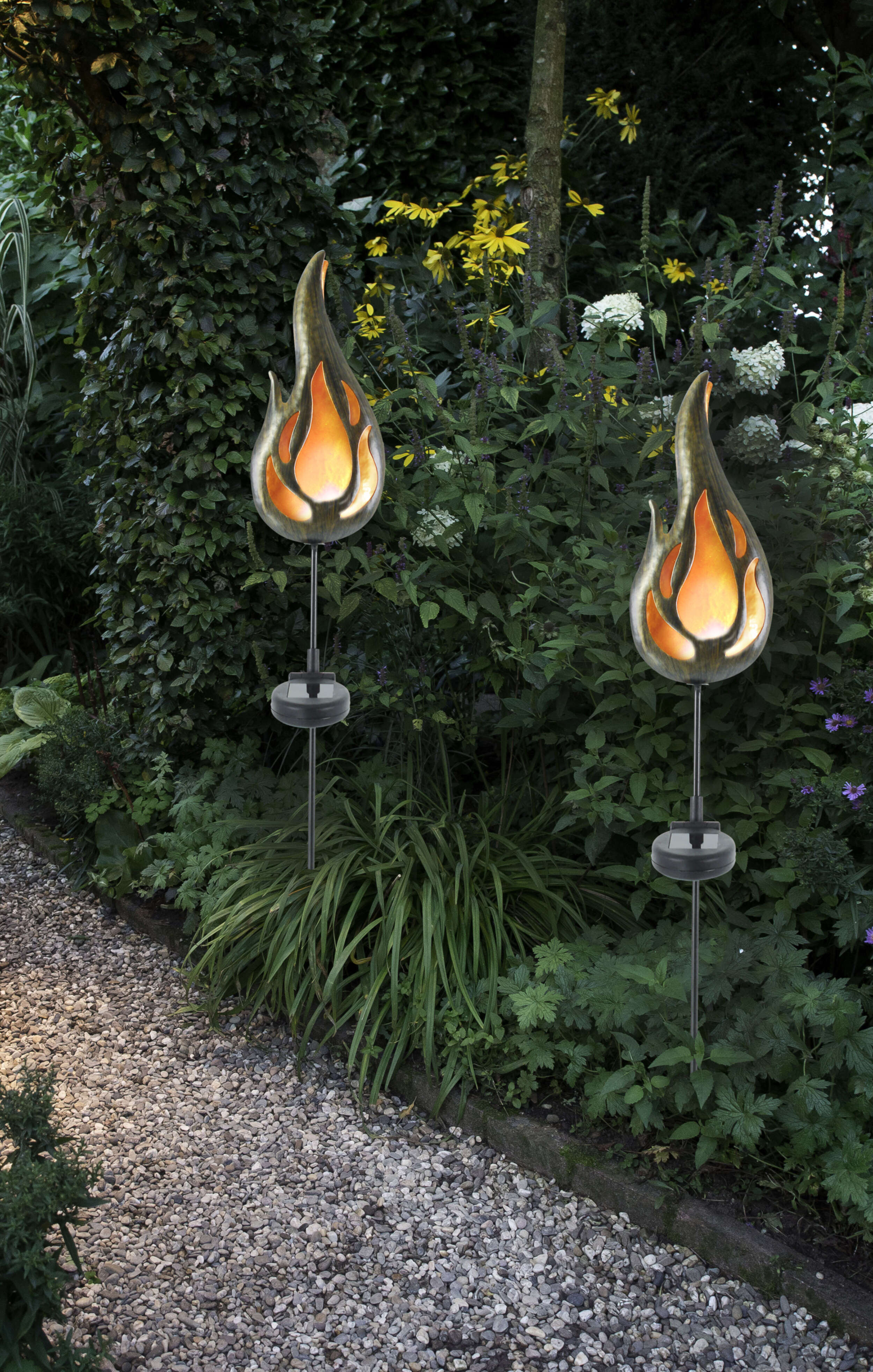 Zwei flammenförmige Außenleuchten in einem Garten. Grünes Laub und Steinweg.