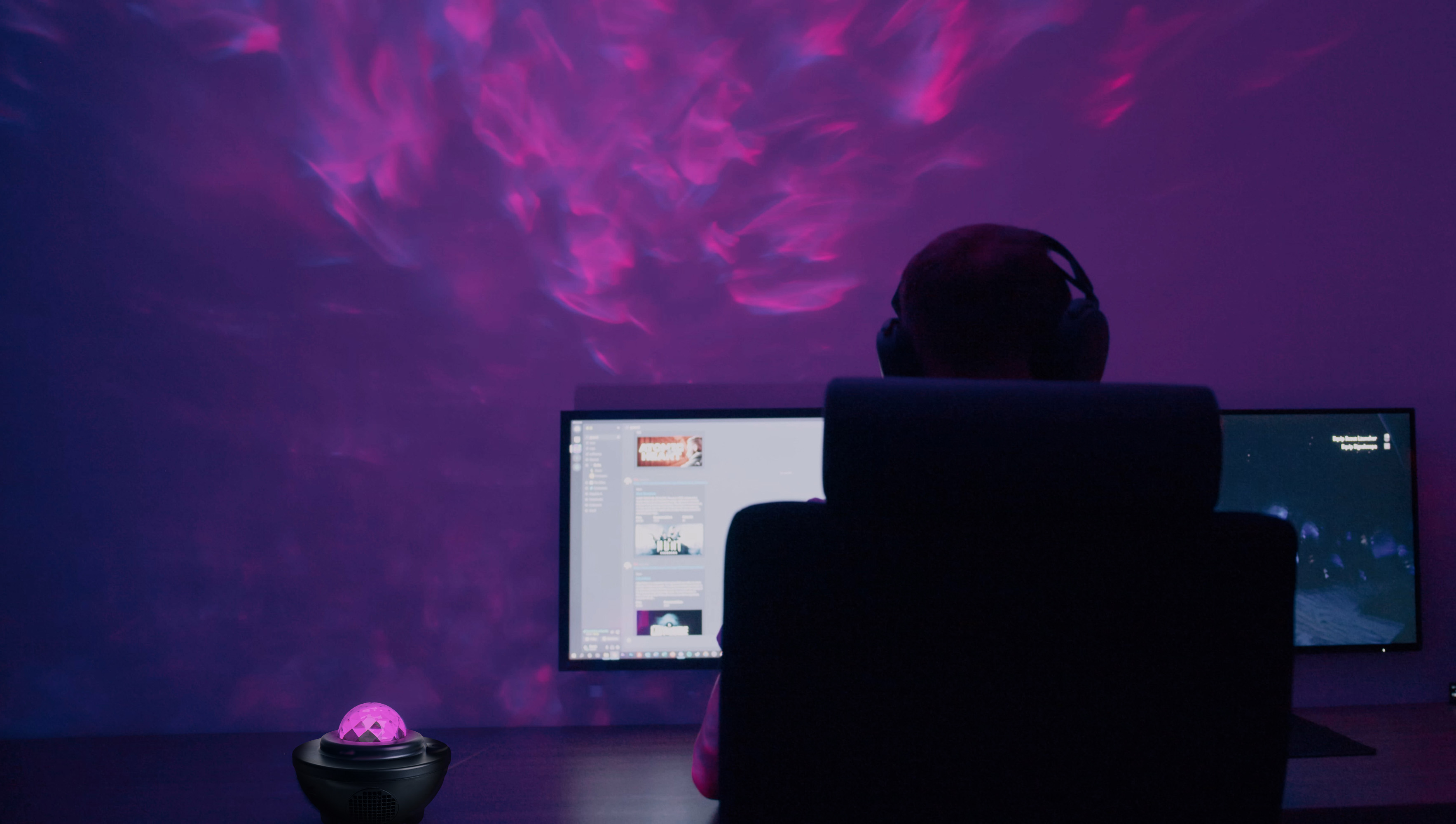 Person mit Kopfhörern, die auf Computerbildschirme blickt, mit lilafarbener, rosa Wolkenprojektion an der Wand.