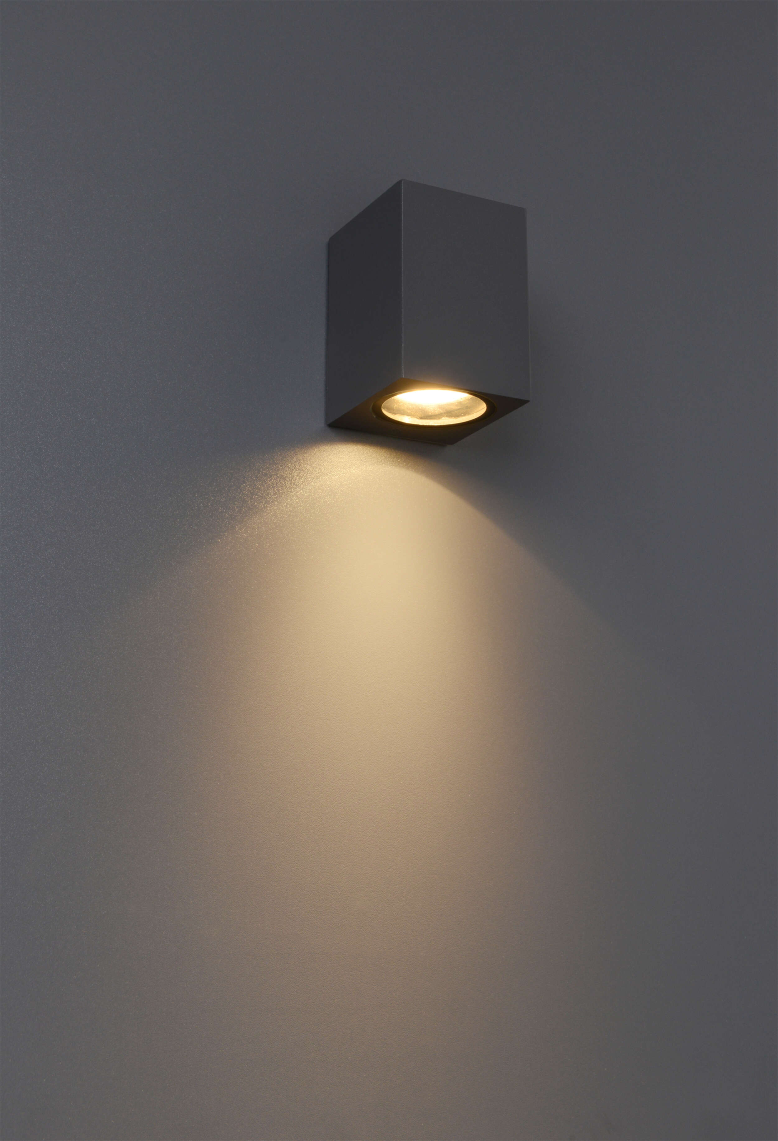 Eine graue, quadratische Leuchte an einer grauen Wand. Sie strahlt ein warmes, nach unten gerichtetes Licht aus.