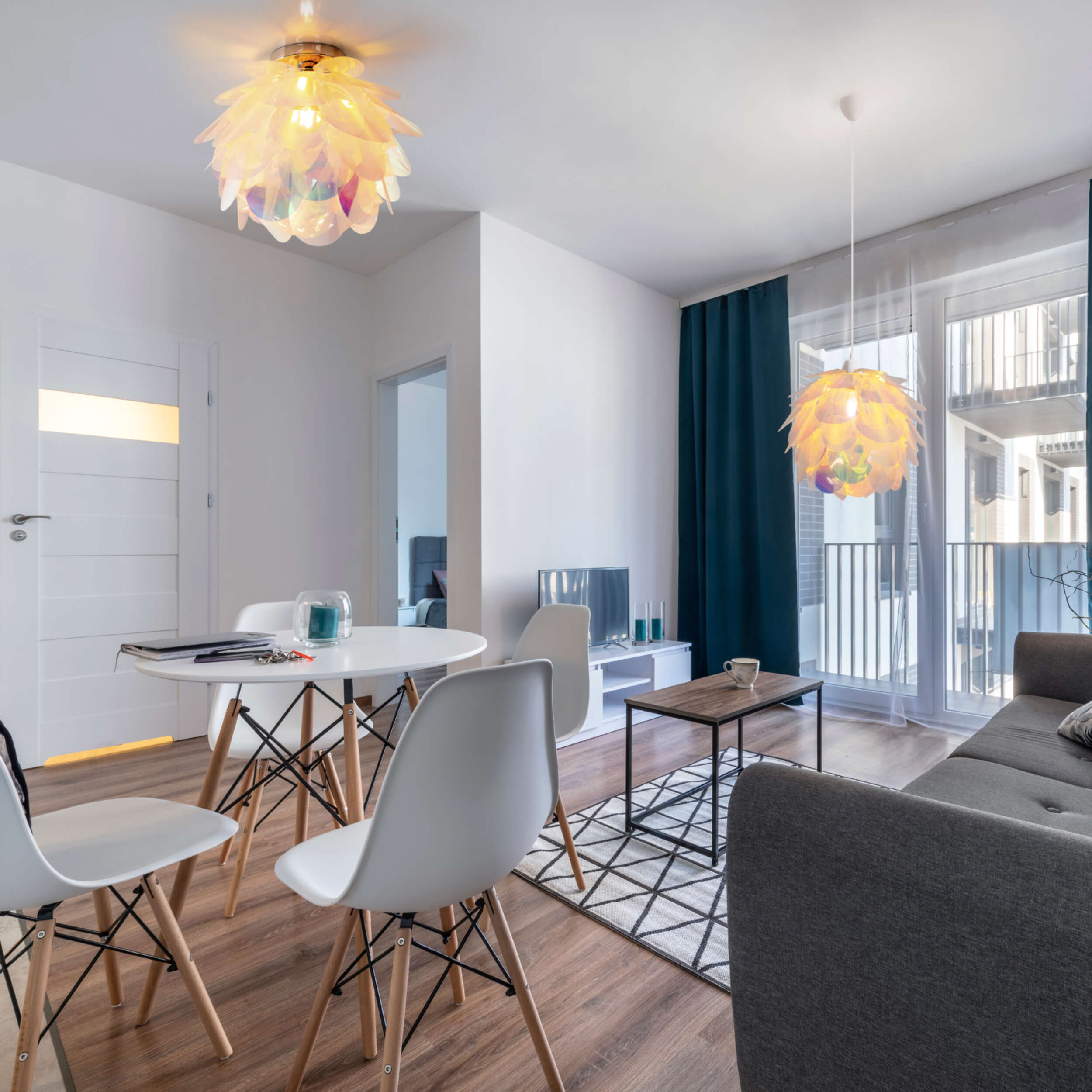 Wohnzimmer mit weißen Wänden, Holzboden, rundem Tisch, weißen Stühlen, Sofa, Teppich und zwei Lampen.
