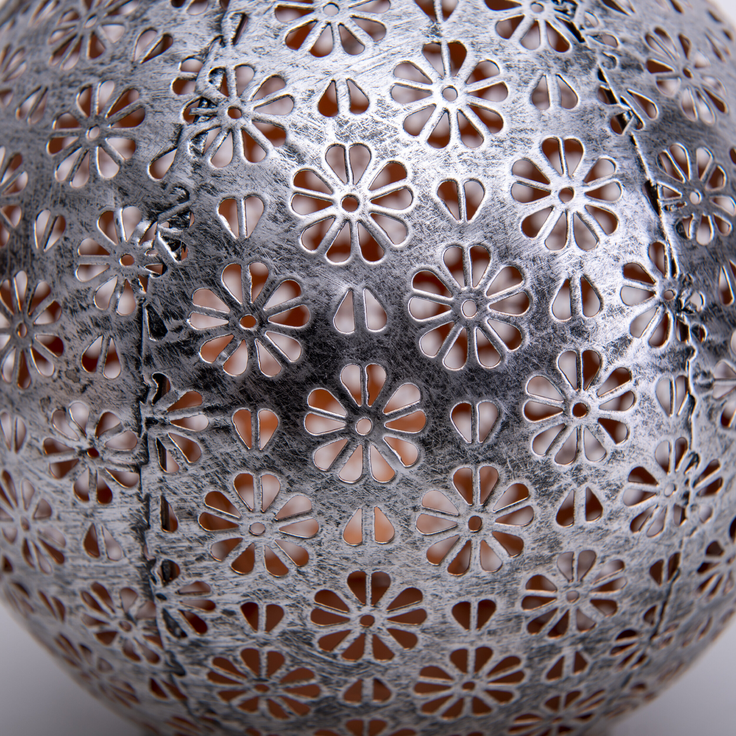 Nahaufnahme einer silbernen dekorativen Kugel mit floralen Ausschnitten. Hintergrund ist hellorange.