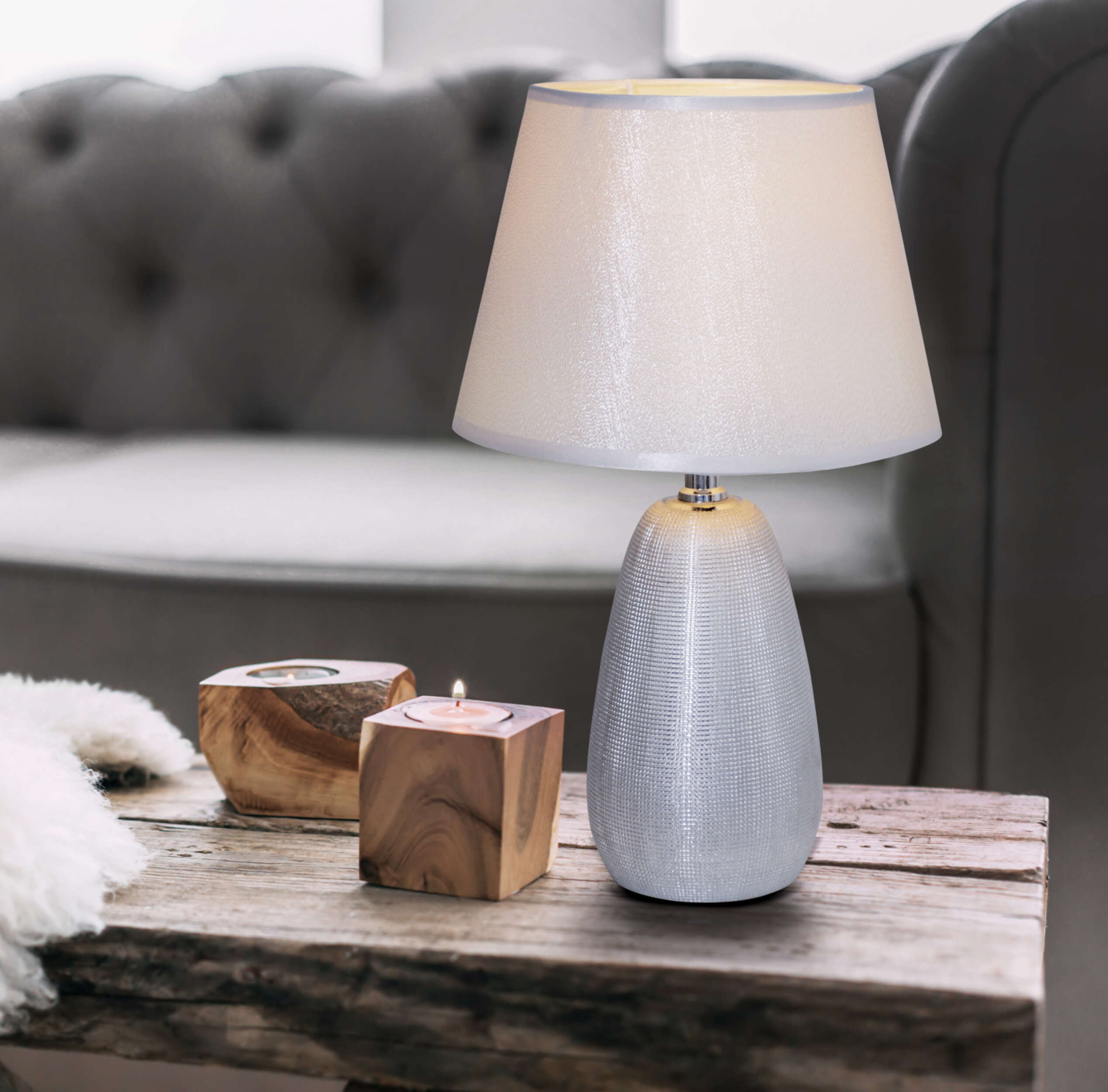 Eine beleuchtete Lampe auf einem Holztisch mit Kerzen und einem Sofa im Hintergrund. Die Lampe hat einen weißen Schirm.
