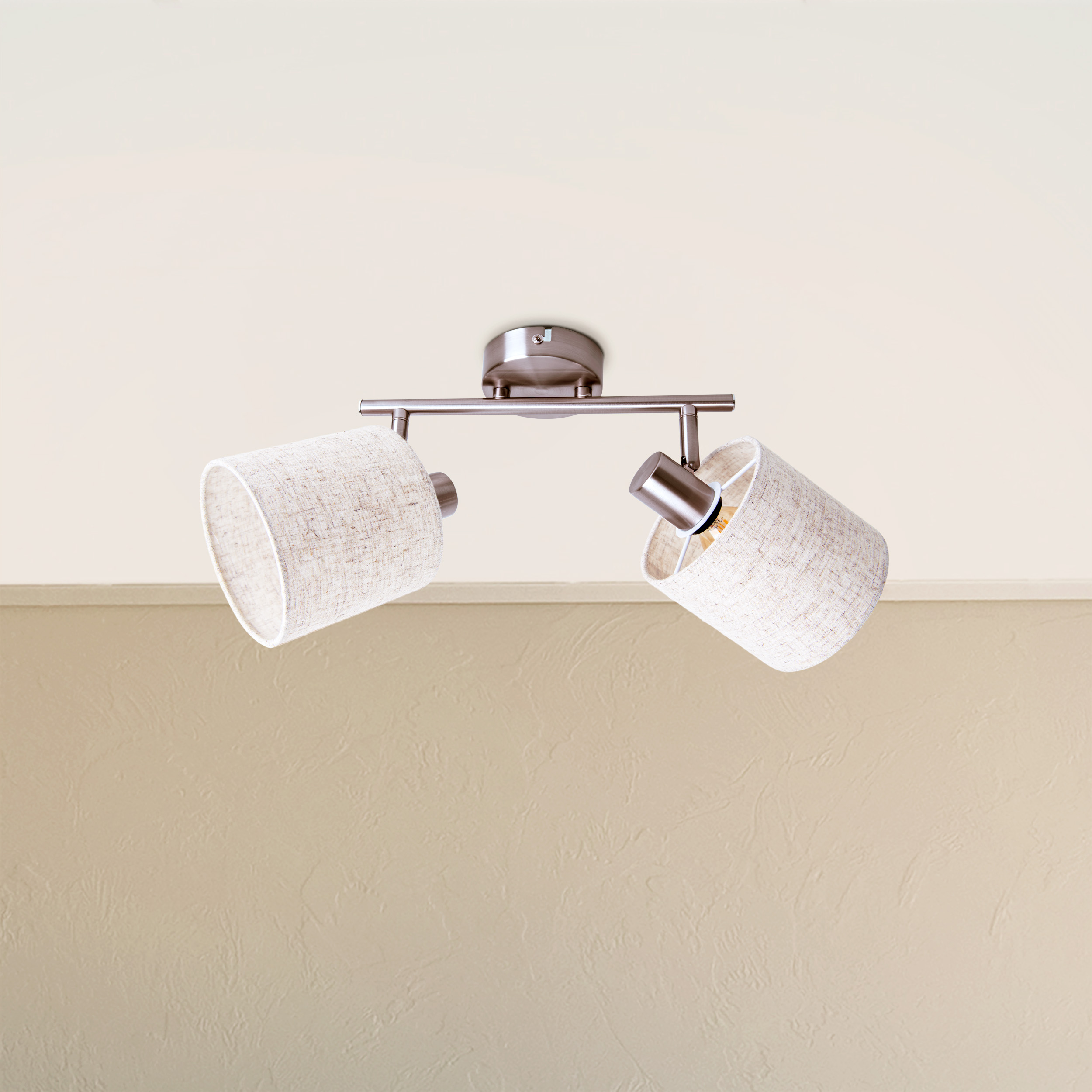 Decken-Spotlight mit zwei Lichtern. Beige Stoffschirme. Silberner Metallrahmen, mit beiger Wand im Hintergrund.