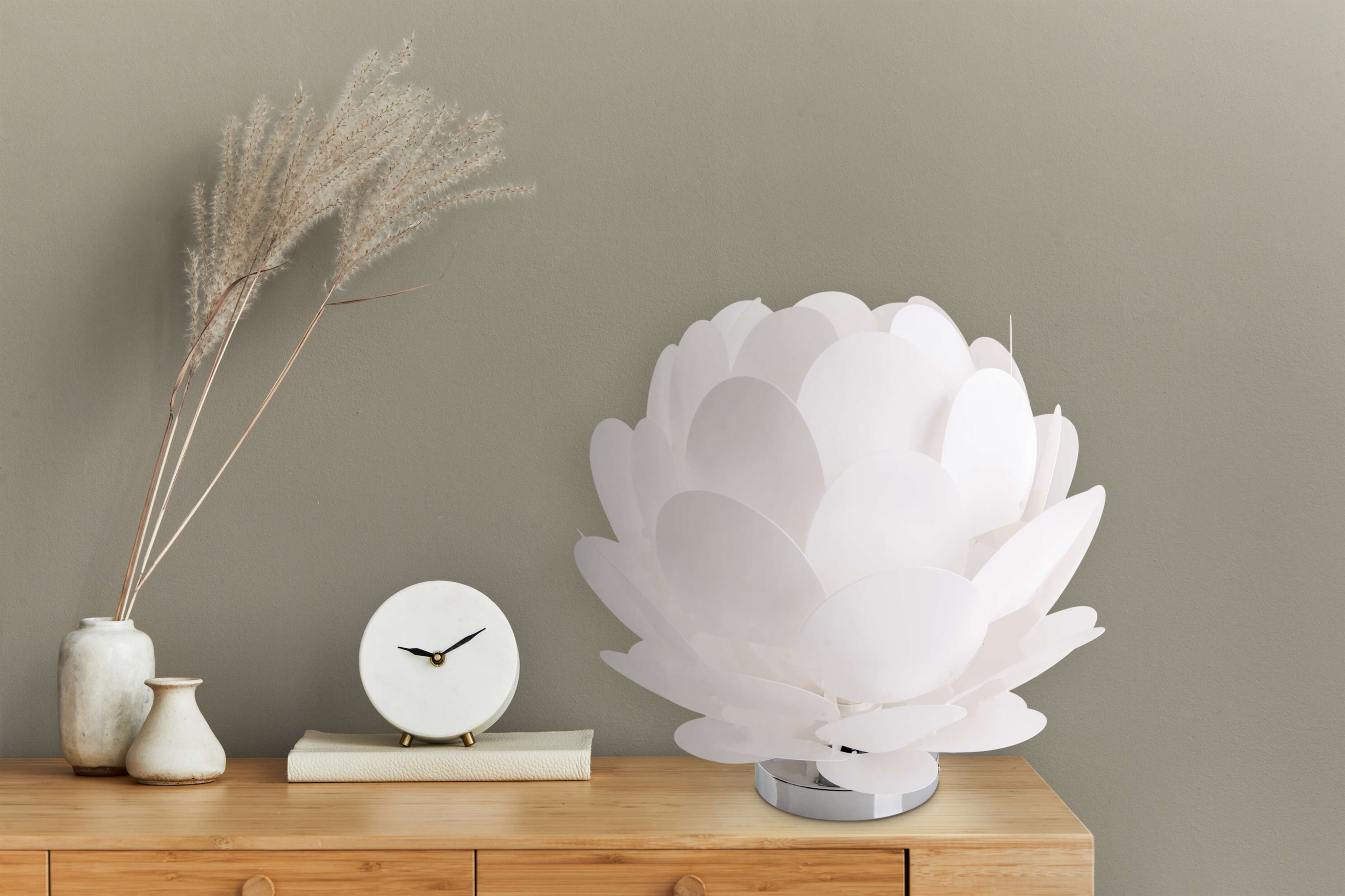 Eine weiße blütenförmige Lampe auf einem Holztisch, mit Uhr und Vase. Im Hintergrund eine neutrale Wand.