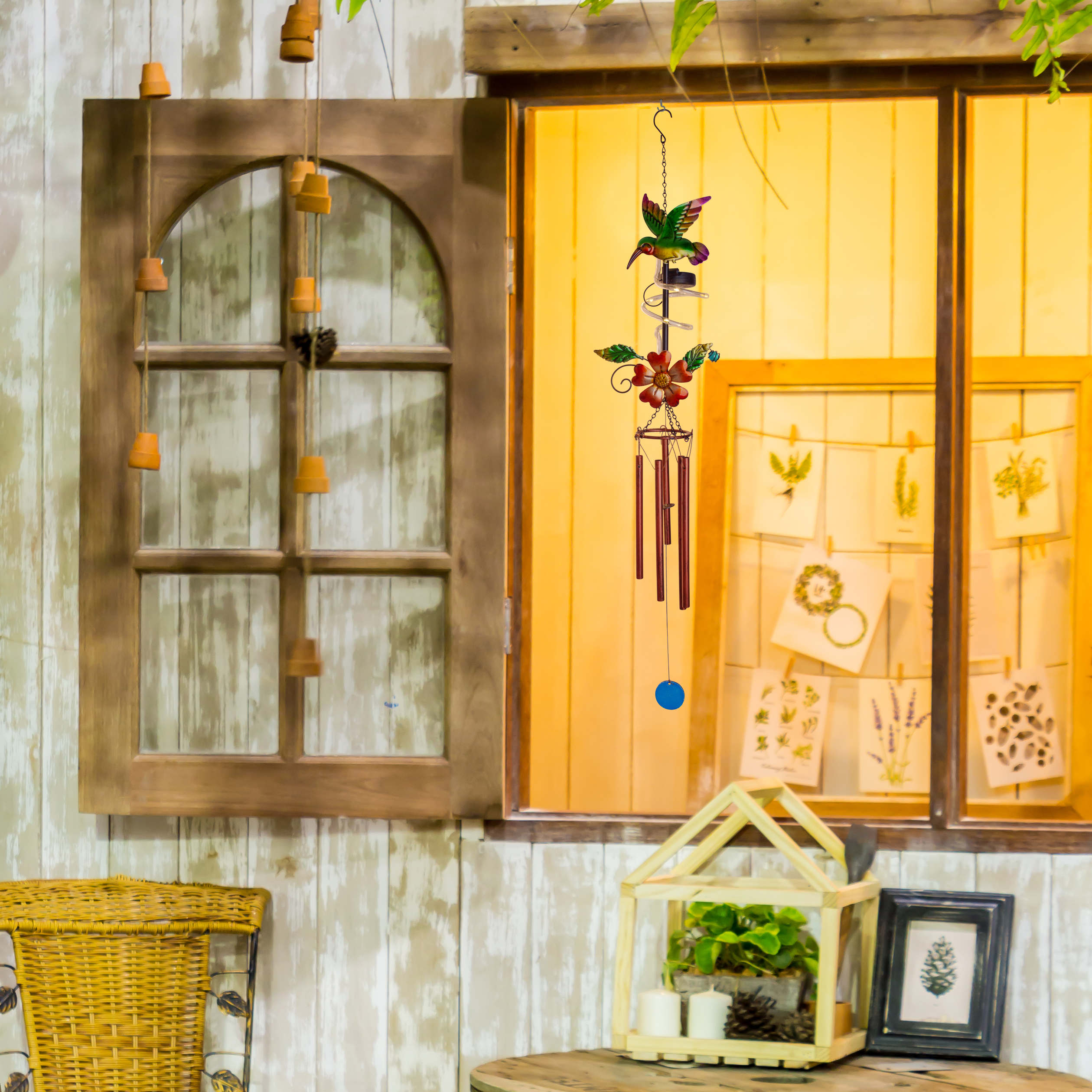 Holzfenster mit Hängedeko und Windspielen. Ein kleiner Tisch mit gerahmtem Bild und hausförmiger Laterne.