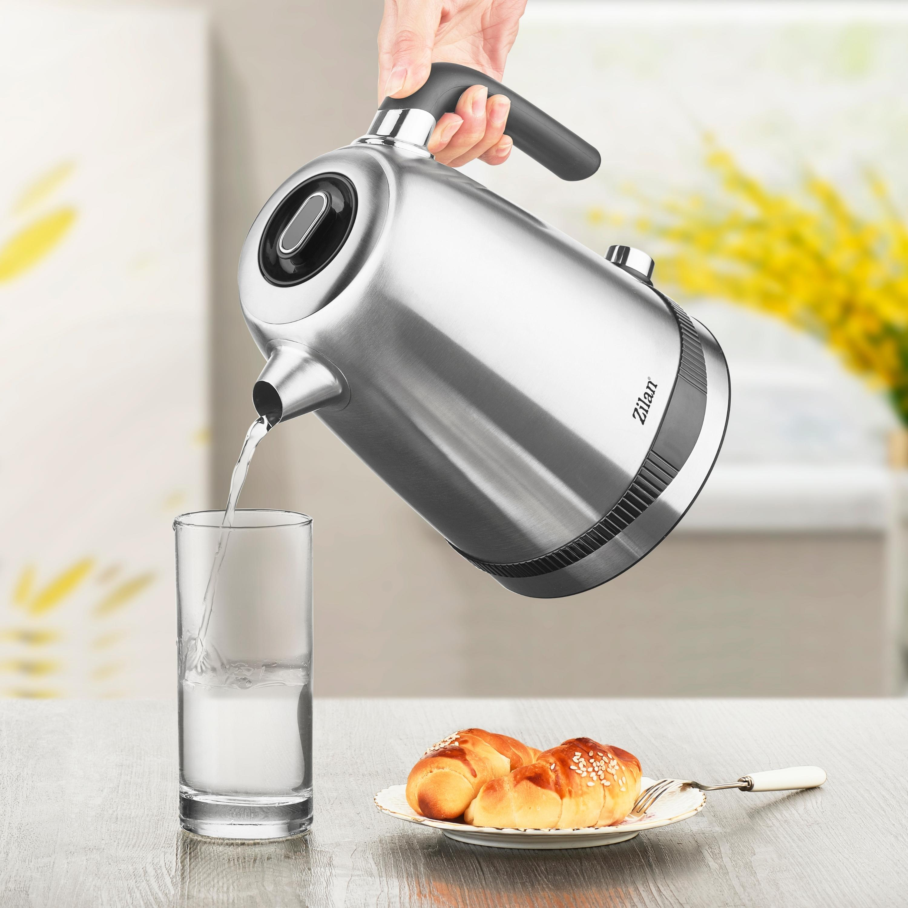 Silberner Wasserkocher gießt Wasser in ein Glas. Teller mit Brötchen auf einem Holztisch. Gelbe Blumen.