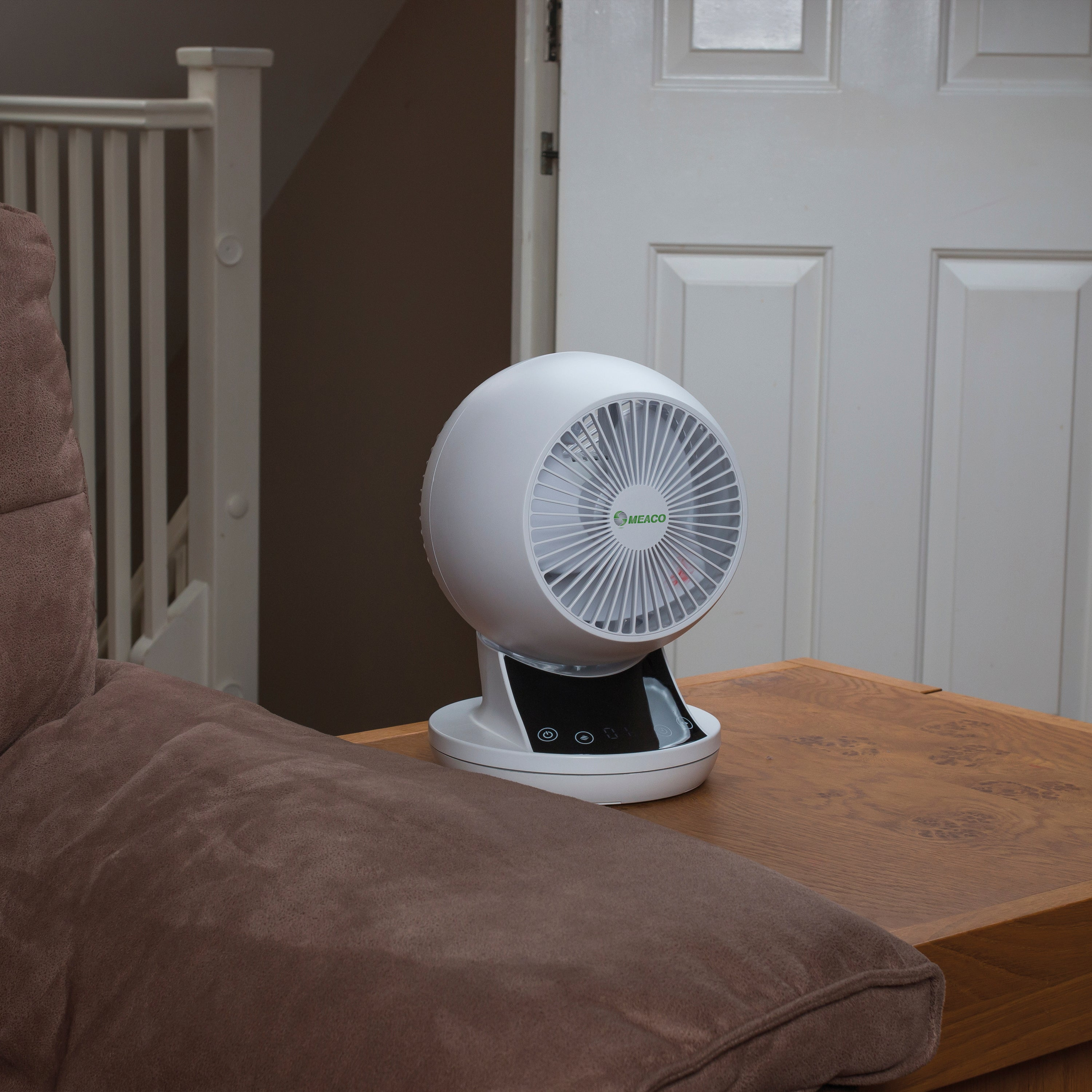 Weißer Ventilator mit kleinem Bildschirm auf einem Tisch, drinnen. Eine Tür und Treppe im Hintergrund.