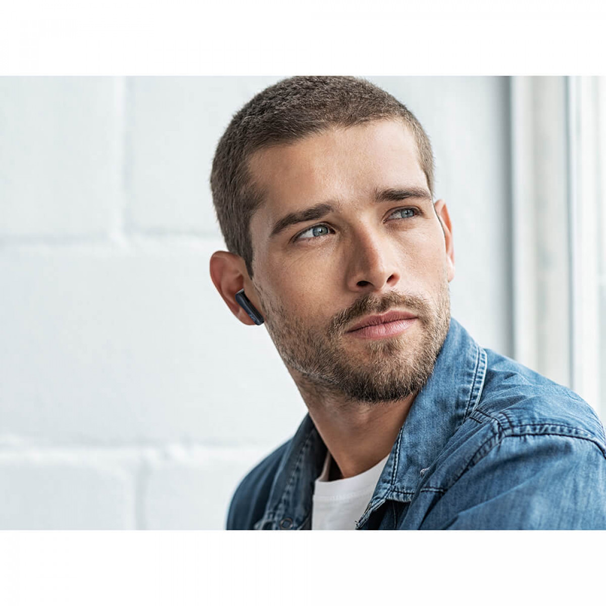 Mężczyzna z brodą, w dżinsowej kurtce, patrzy w bok. Ma niebieską słuchawkę w lewym uchu.