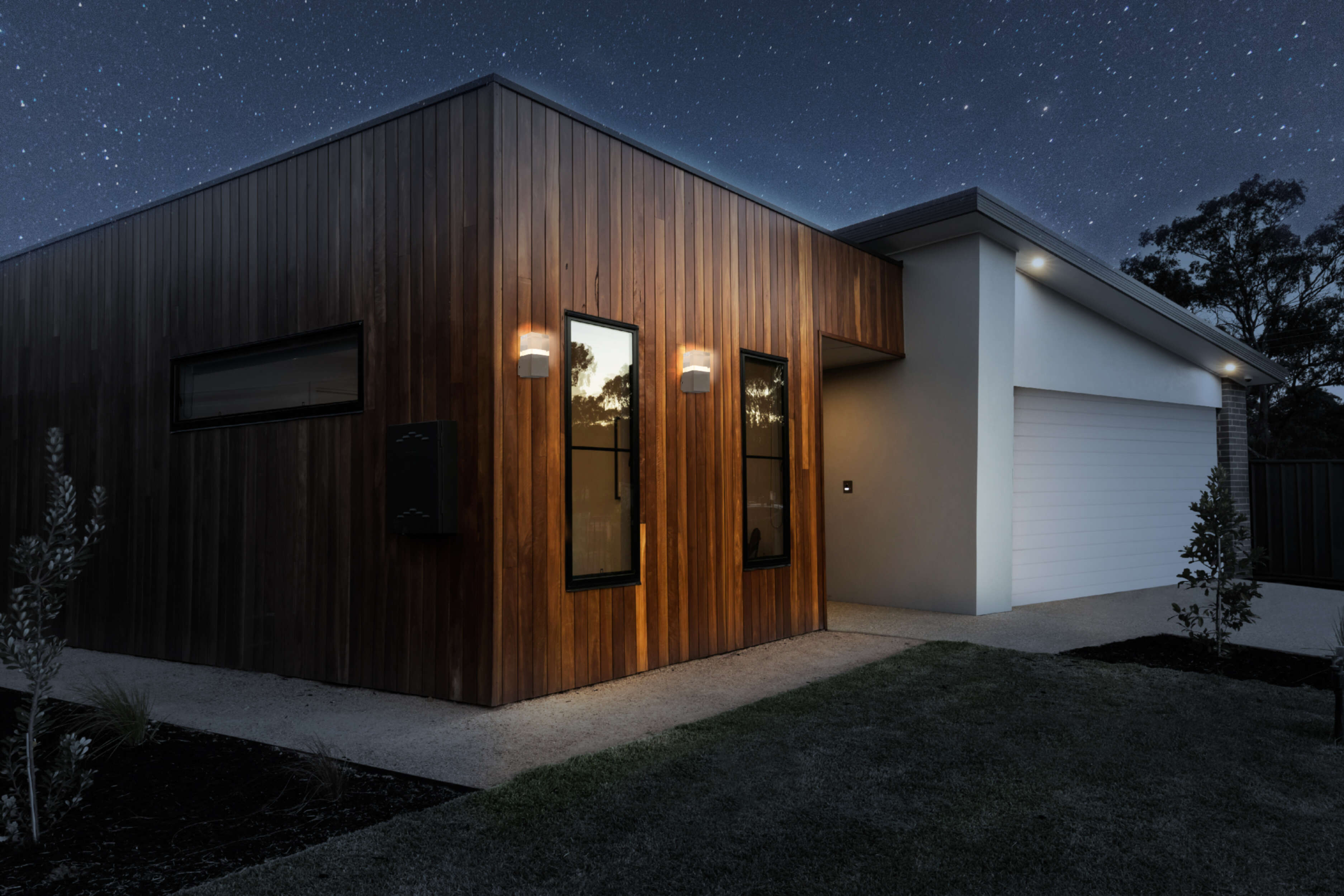 Modernes Haus bei Nacht. Holzfassade, Lichter an, Sternenhimmel im Hintergrund.