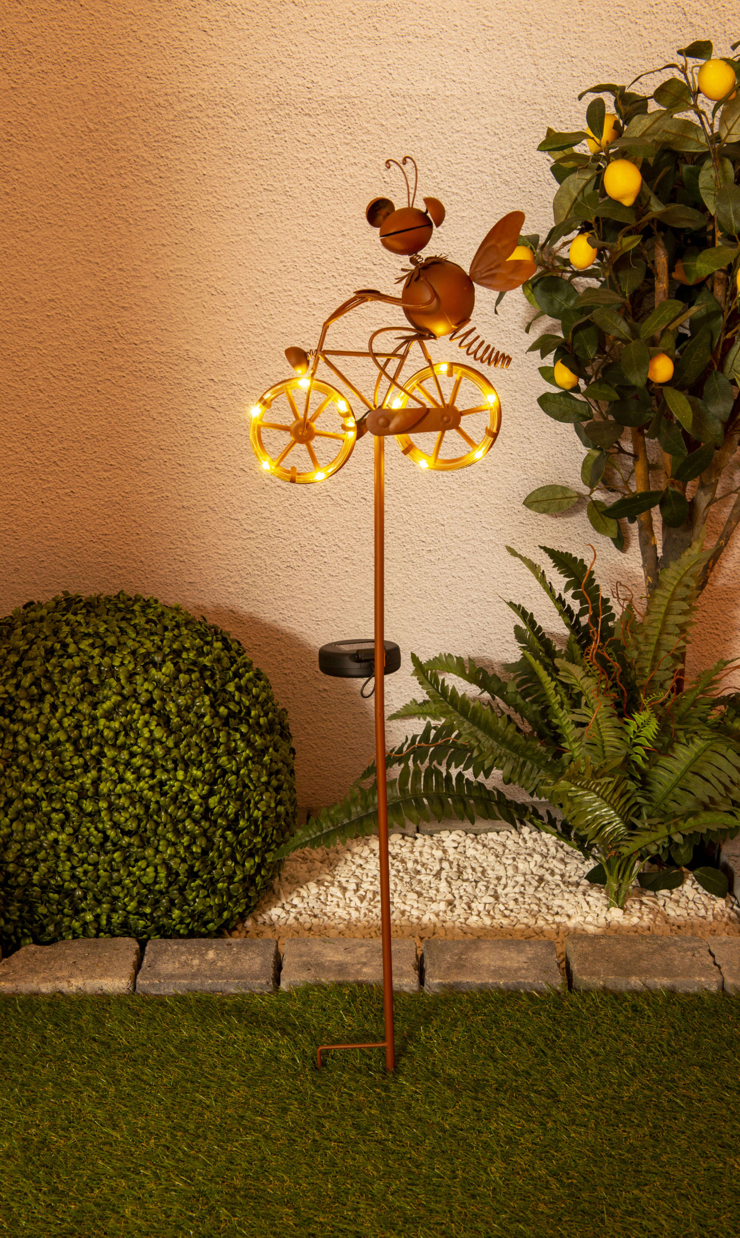 Metallbiene auf Fahrrad mit leuchtenden Rädern im Garten, in der Nähe von Busch und Zitronenbaum.