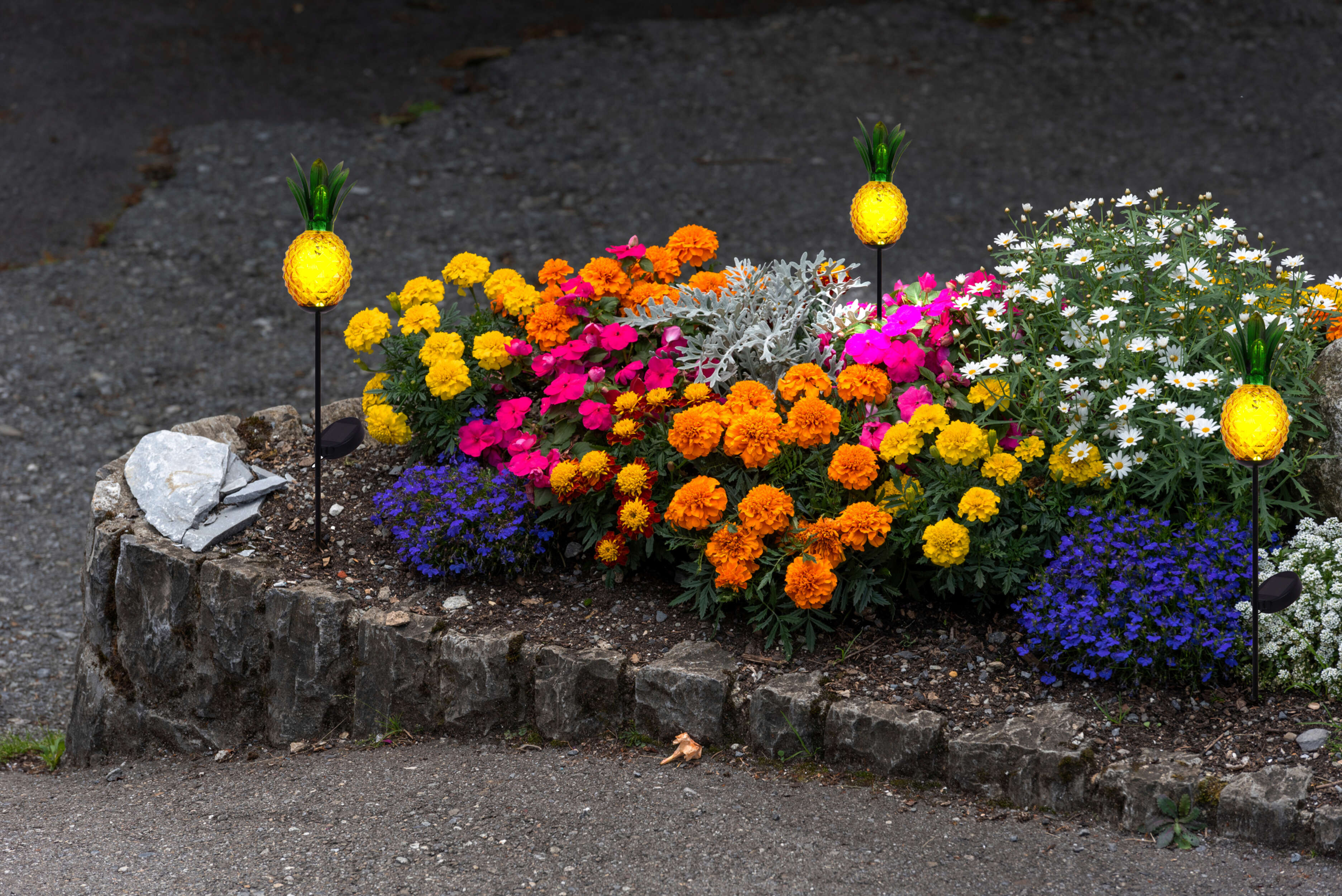 Ein Blumenbeet mit bunten Blumen und ananasförmigen Solarleuchten mit schwarzen Pfählen.