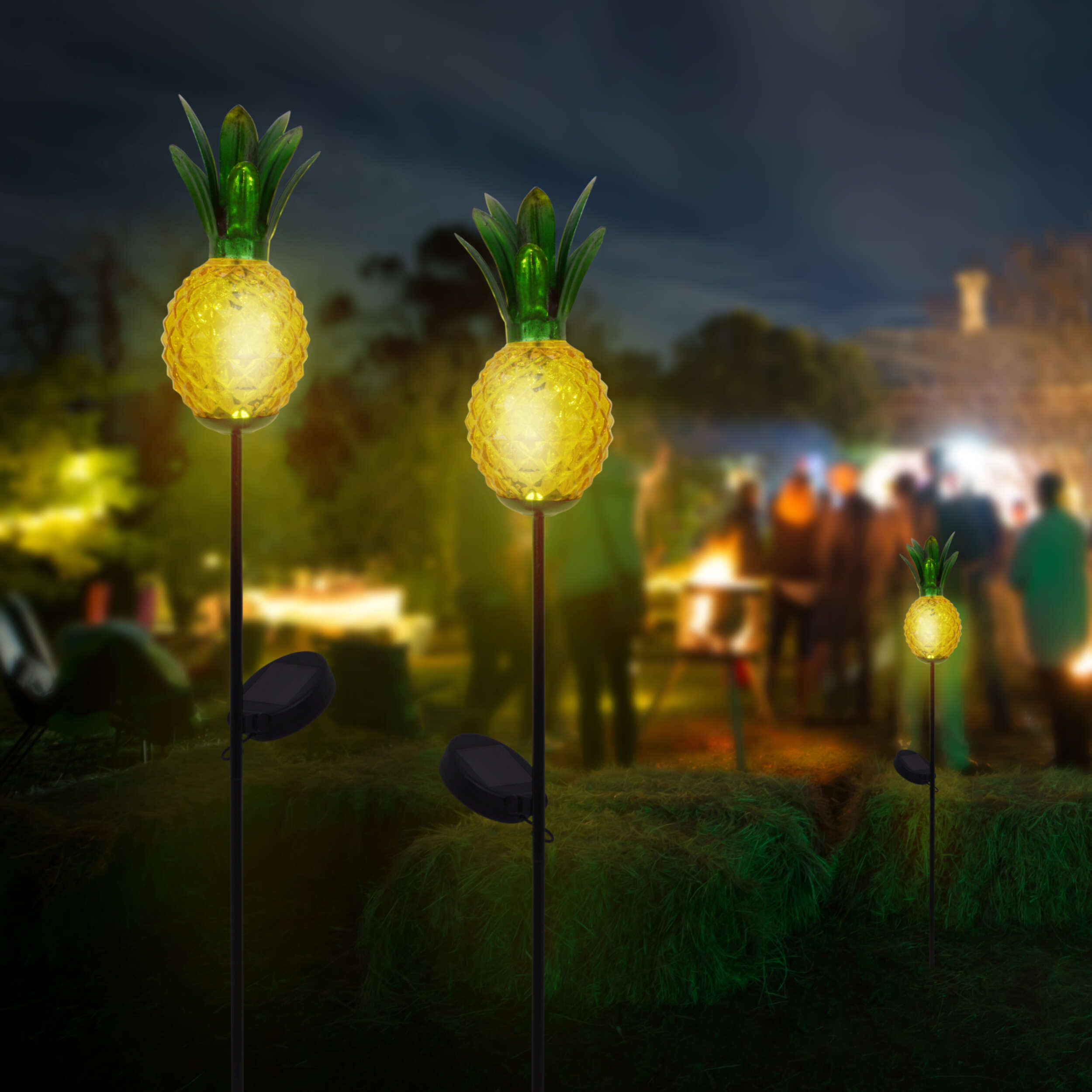Drei solarbetriebene Lichter in Ananasform auf einem Feld. Verschwommener Hintergrund mit Menschen und Feuer.