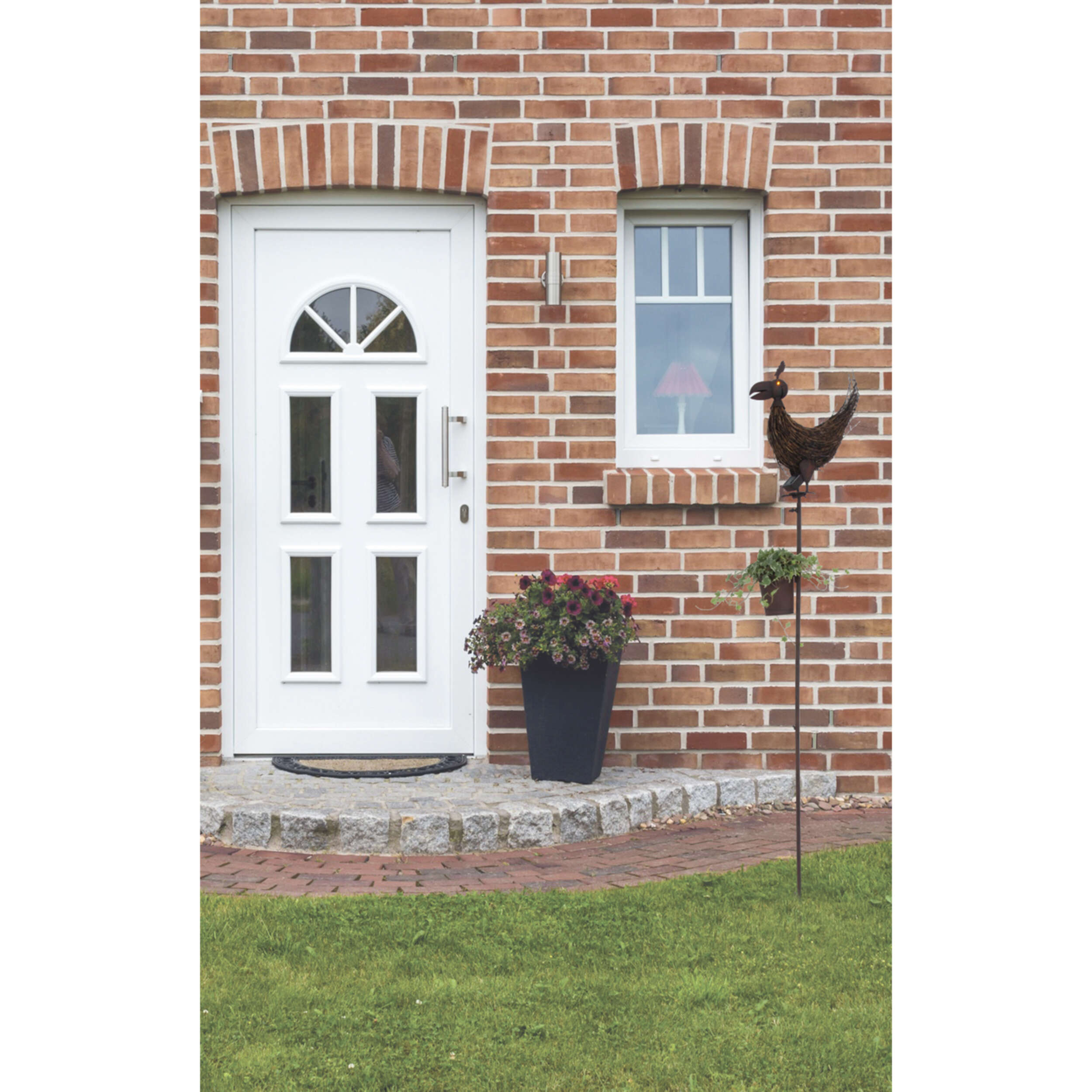 Ziegelmauer mit weißer Tür und Fenster. Eine Vogel-Windfahne und Blumen sind davor.