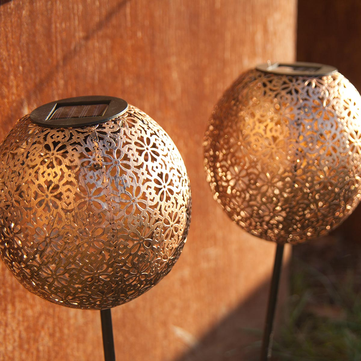 Zwei kugelförmige, goldene Solar-Gartenleuchten mit dekorativen Ausschnitten, auf braunem Hintergrund.