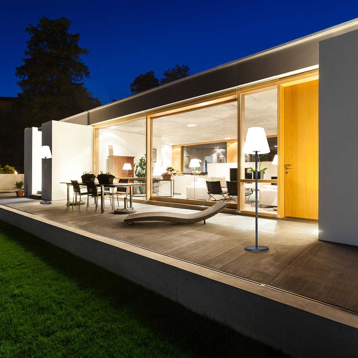 Moderne Terrassenszene bei Nacht. Lichter beleuchten den Raum mit Esstisch und Liegestuhl.