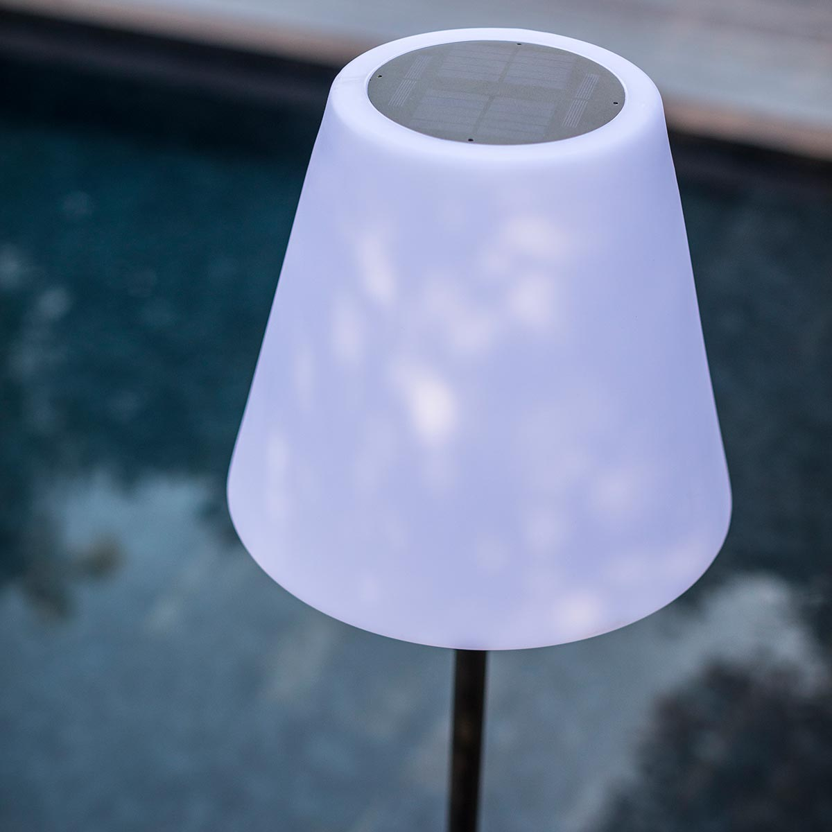 Weiße Solarlampe mit konischem Schirm, leuchtend. Der Hintergrund ist ein verschwommener Pool.