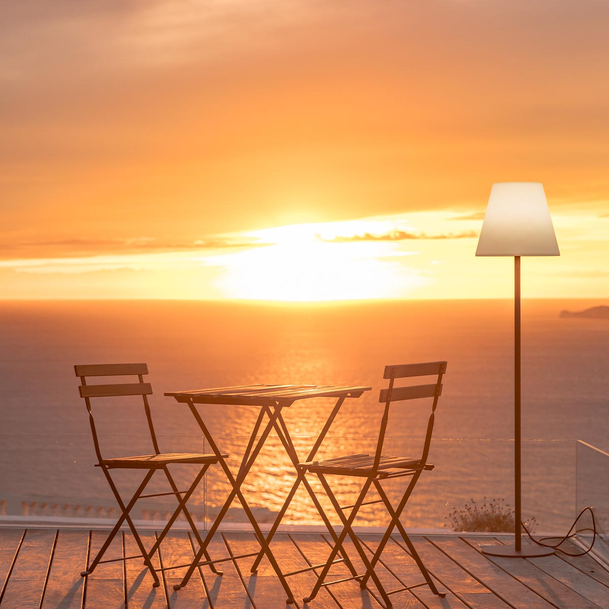 Sonnenuntergangsszene mit Tisch und Stühlen auf einem Deck, Lampe und Meerblick.