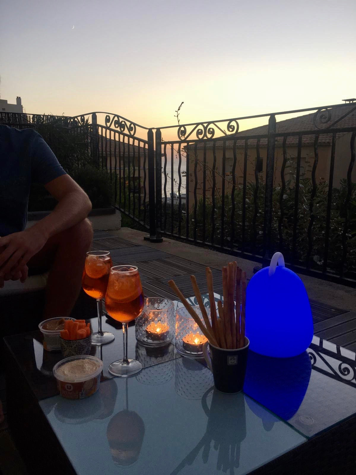 Außenbereich. Zwei Aperol Spritz, Snacks, Kerzen und ein blaues Licht. Sonnenuntergang im Hintergrund.