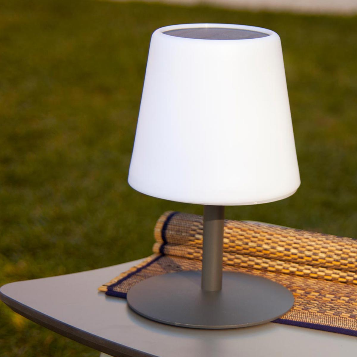 Weiße solarbetriebene Lampe mit grauem Sockel auf einem Tisch mit gewebter Matte.