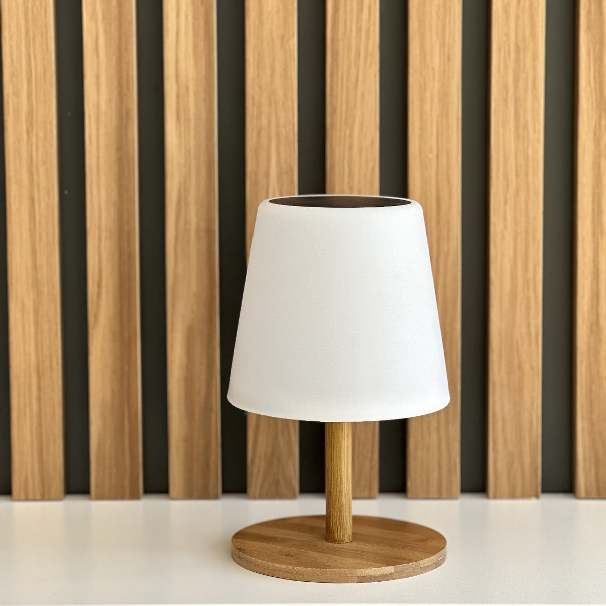 Weiße Tischlampe mit Holzfuß und Schirm. Im Hintergrund eine Wand mit Holzlatten.