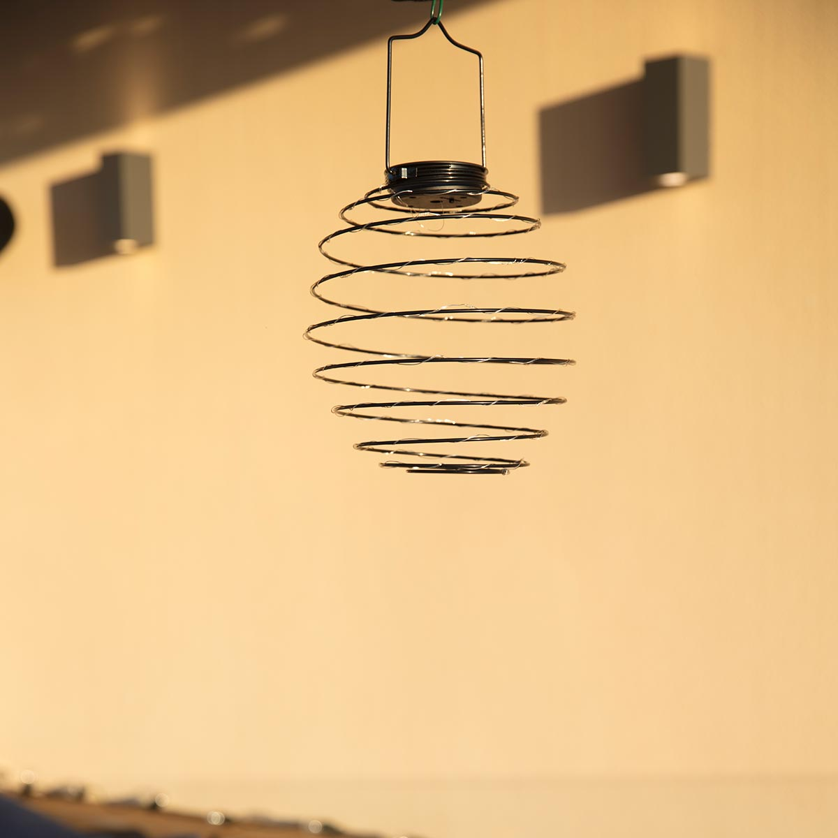 Eine metallene Spiral-Hängeleuchte vor einer beigefarbenen Wand, mit Sonnenlicht.