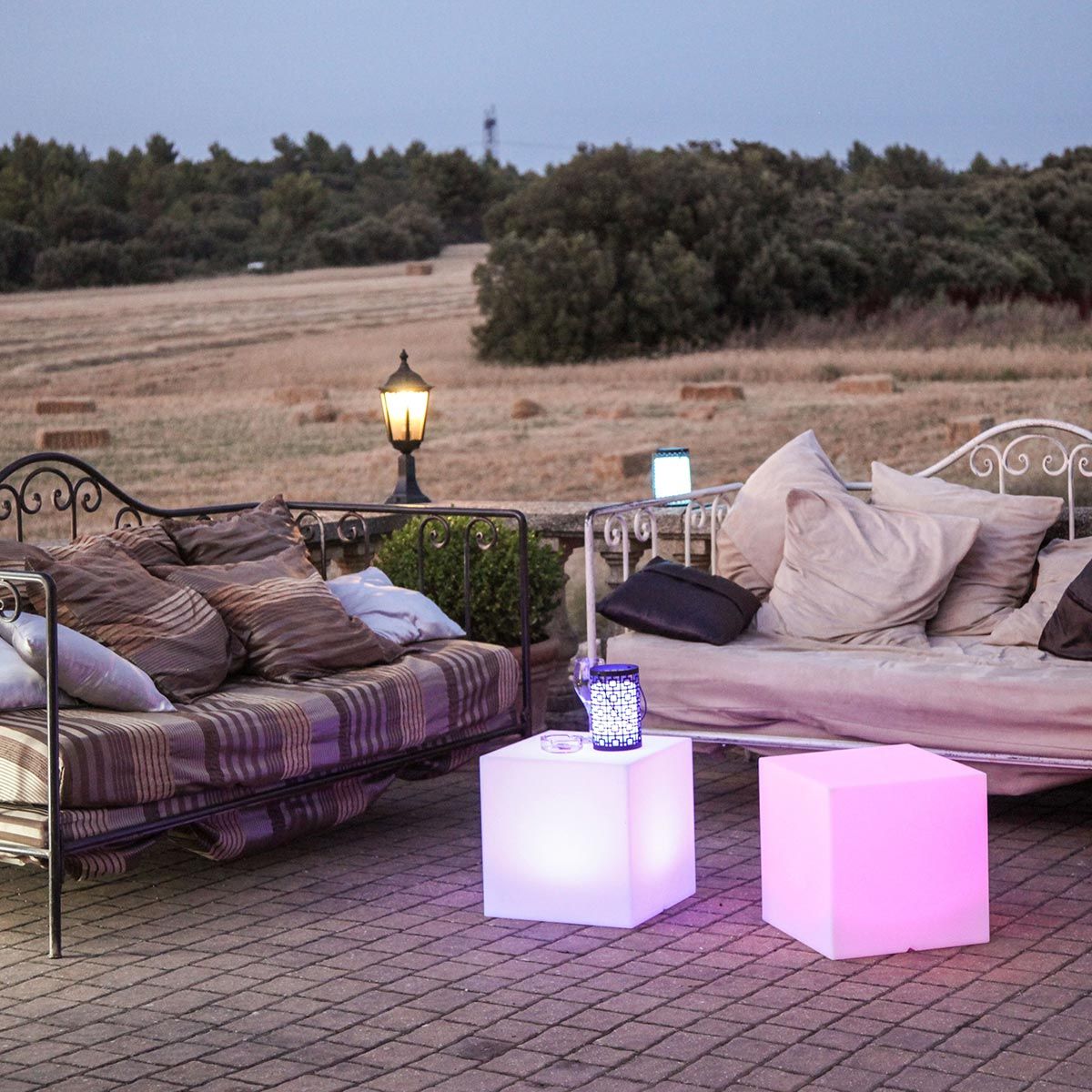 Szene im Freien mit zwei beleuchteten, leuchtenden Würfeltischen. Sofas auf einer Ziegelterrasse, Felder und Bäume im Hintergrund.
