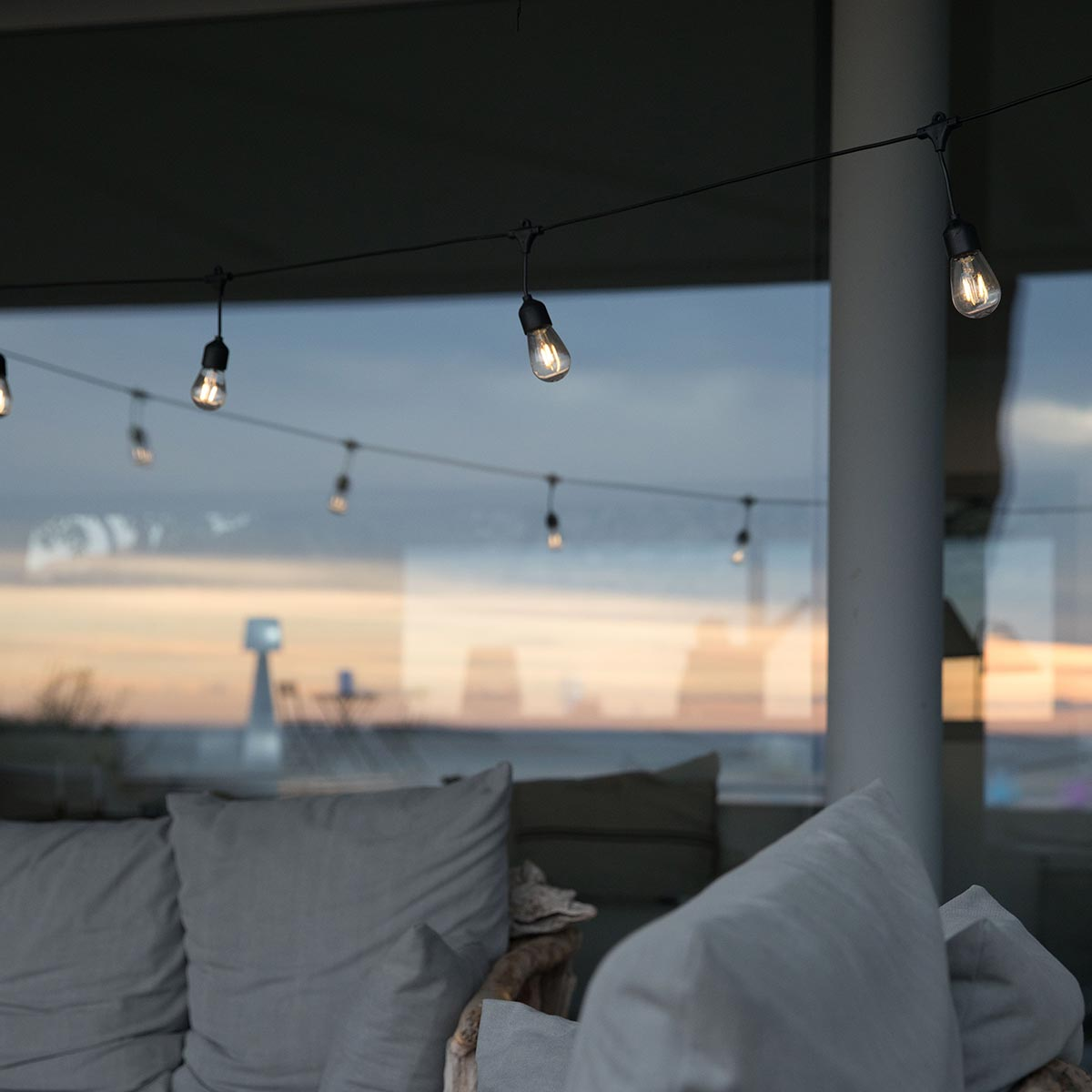 String lights with small bulbs hanging against a window with a sunset view and some outdoor seating.
