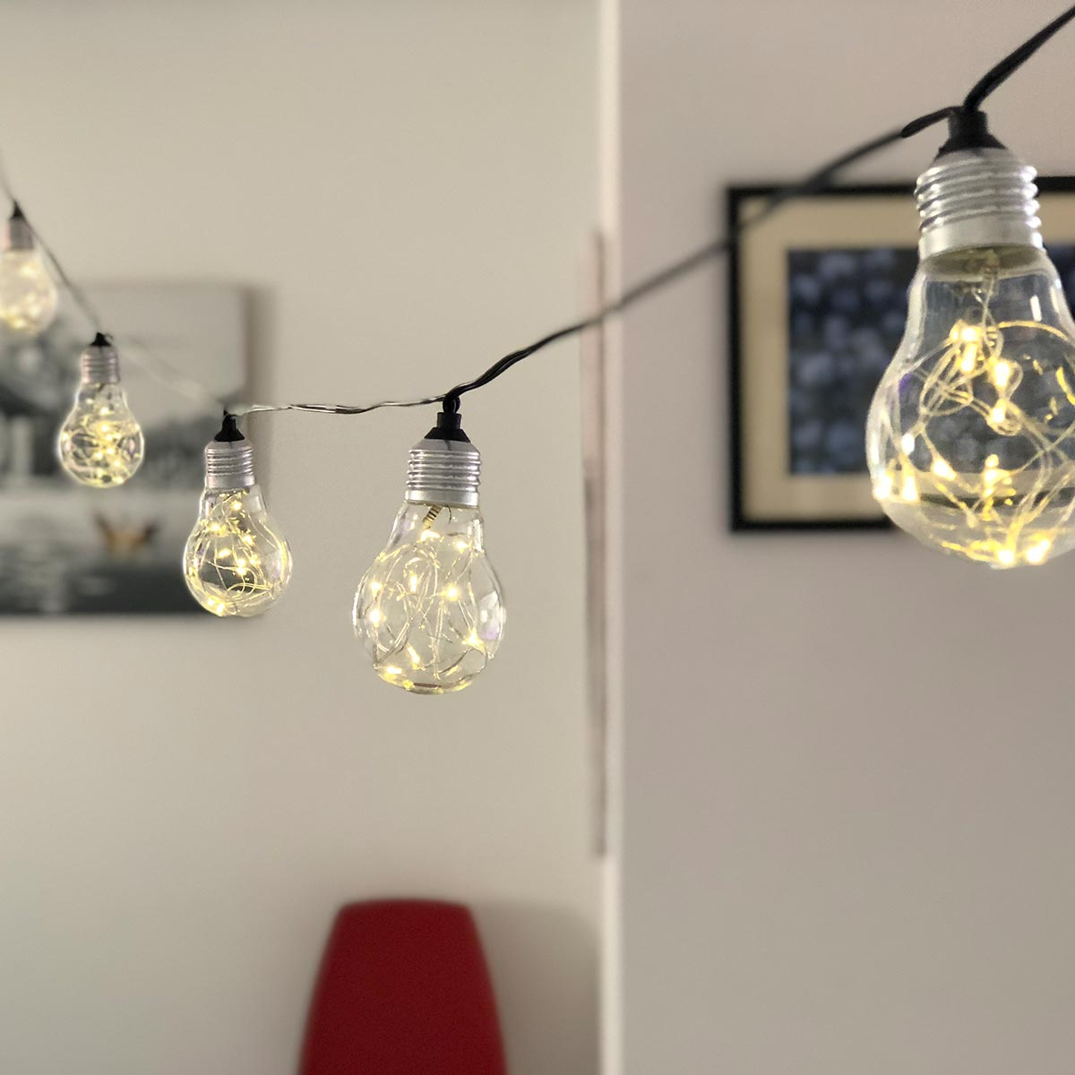 Lichterketten mit klaren Glühbirnen hängen vor einer weißen Wand und einem gerahmten Bild.