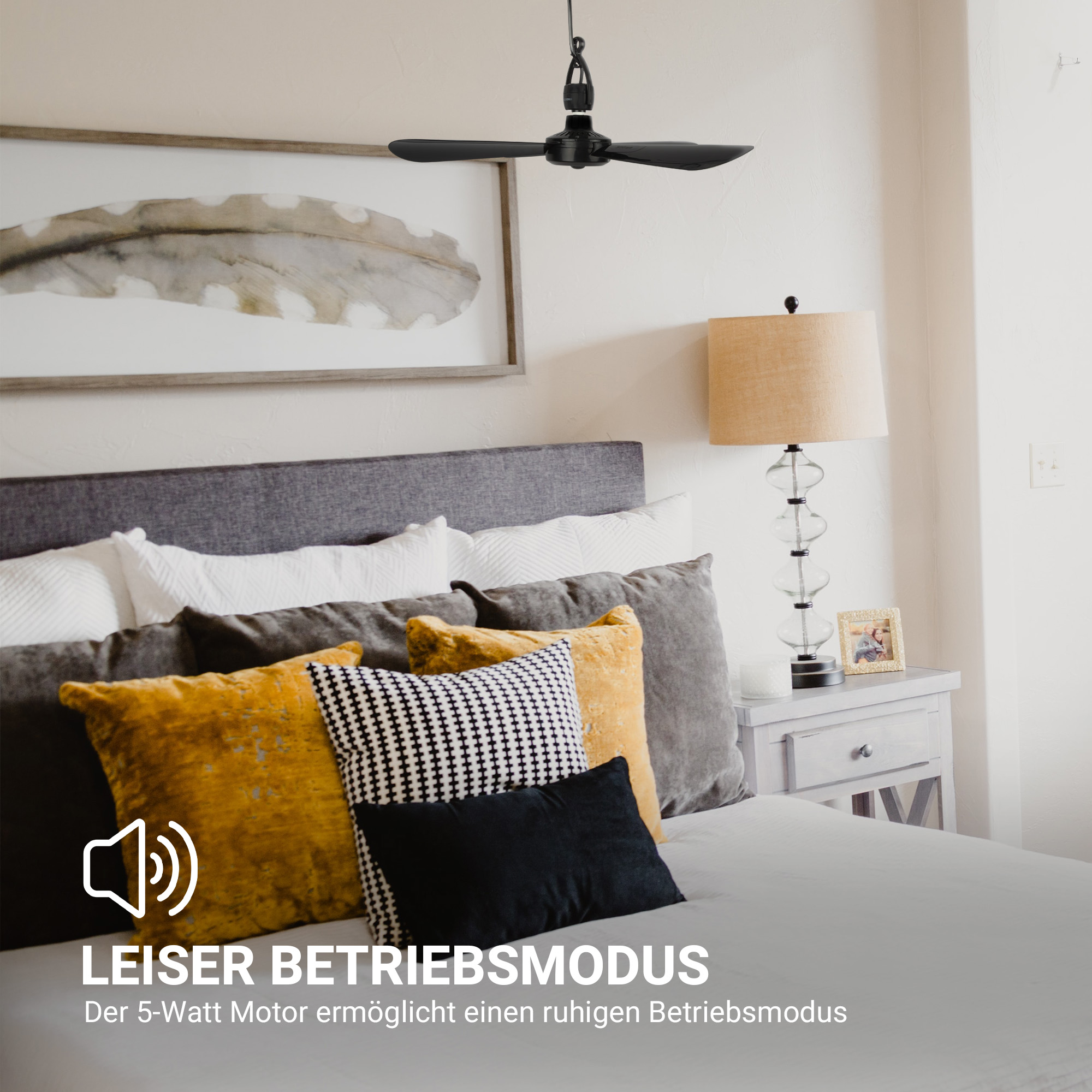 Ein Schlafzimmer mit Deckenventilator, Bett mit Kissen, Lampe und Bilderrahmen an der Wand.