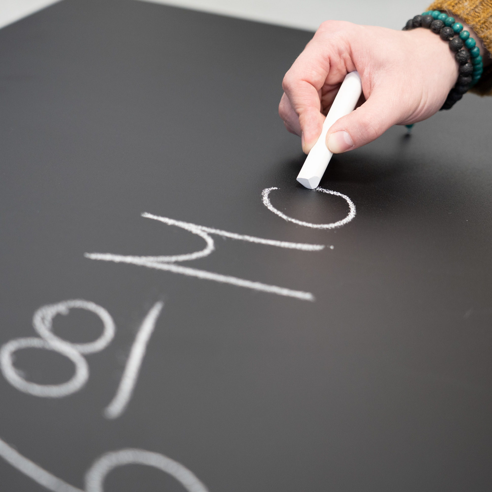 Eine Hand, die mit weißer Kreide auf eine Tafel schreibt und die Zahlen 81 und 76 schreibt.