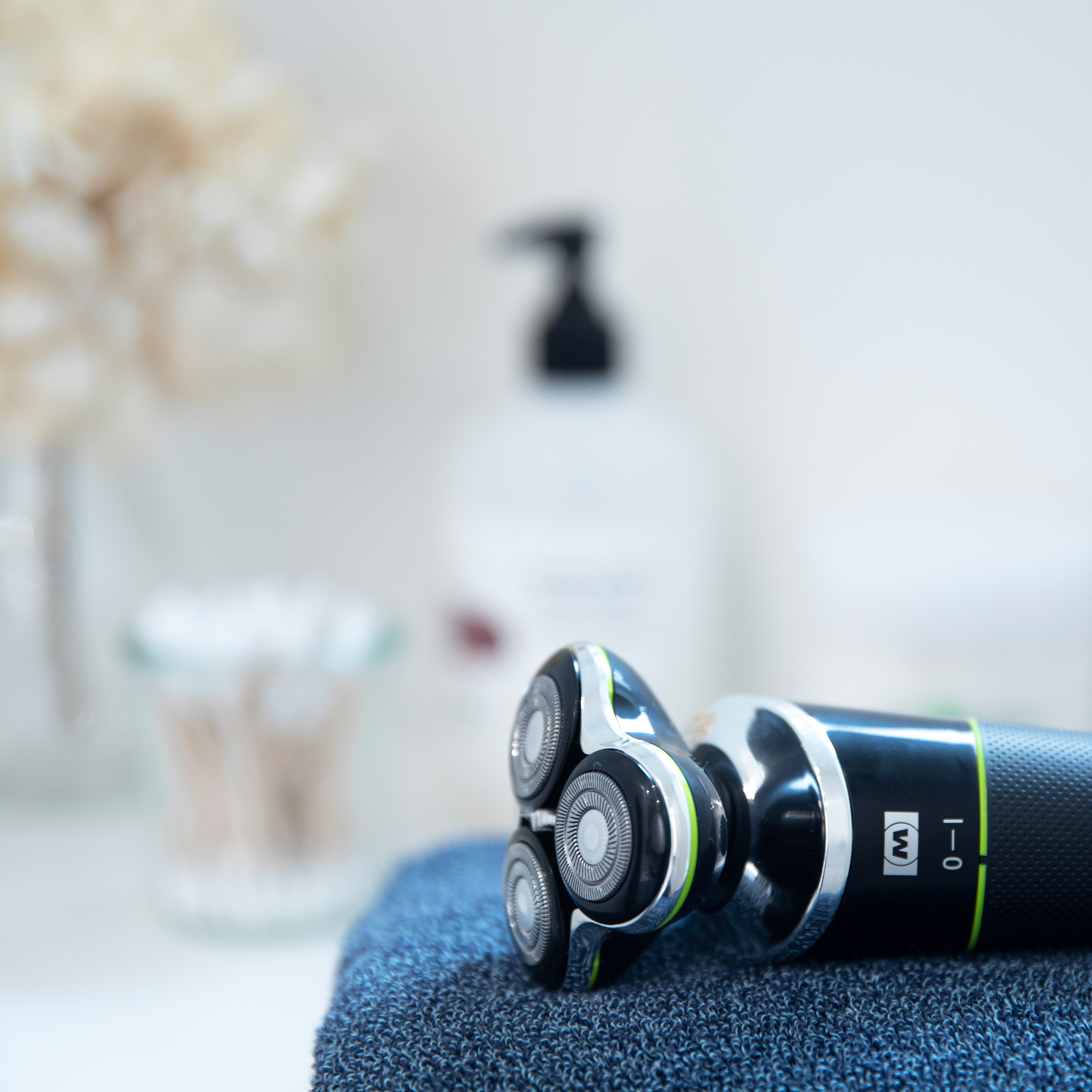 Golarka elektryczna na niebieskim ręczniku. Rozmyte tło z widocznymi kosmetykami.