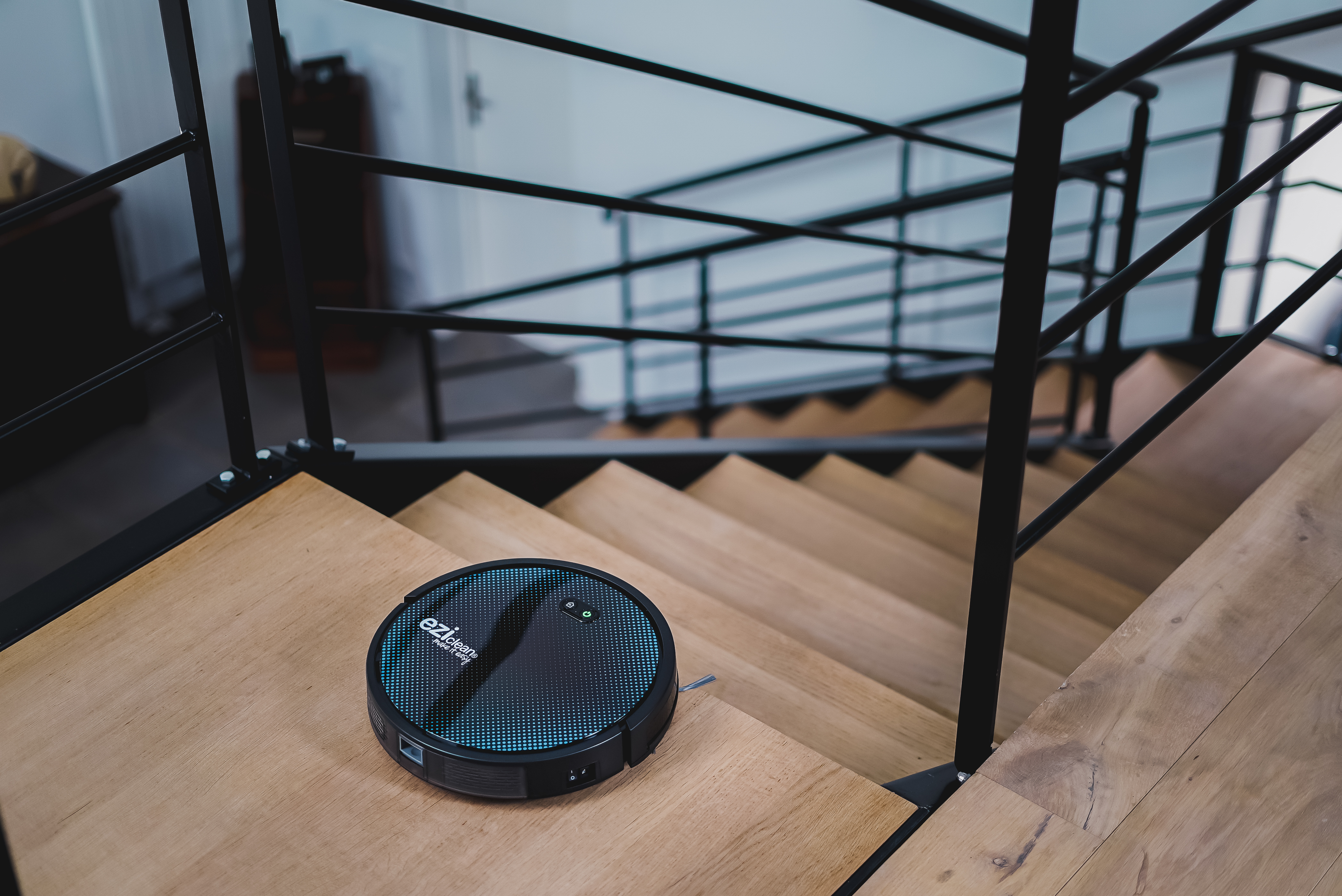 Aspiradora robot en escaleras de madera, cerca de una barandilla negra.