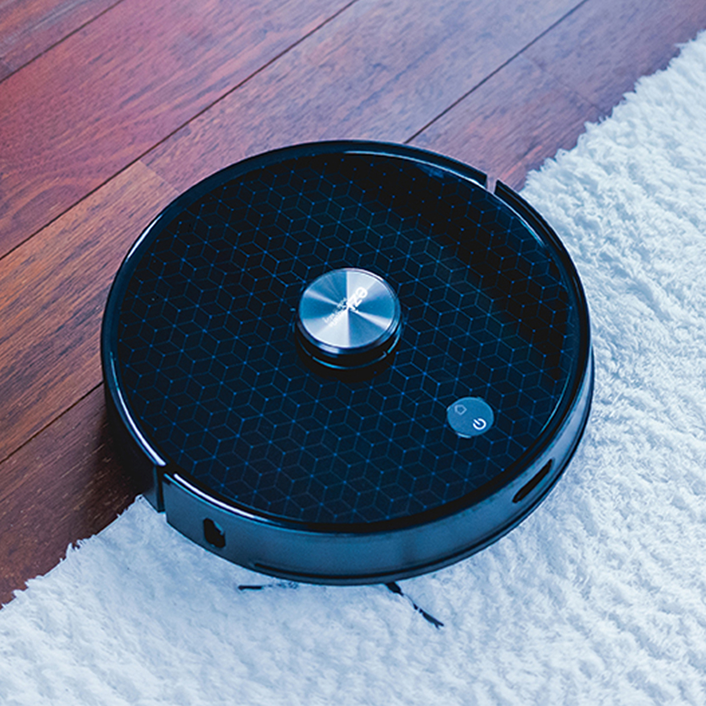 Ein schwarzer, runder Roboter-Staubsauger auf einem Holzboden und einem weißen Teppich. Der Power-Knopf leuchtet.