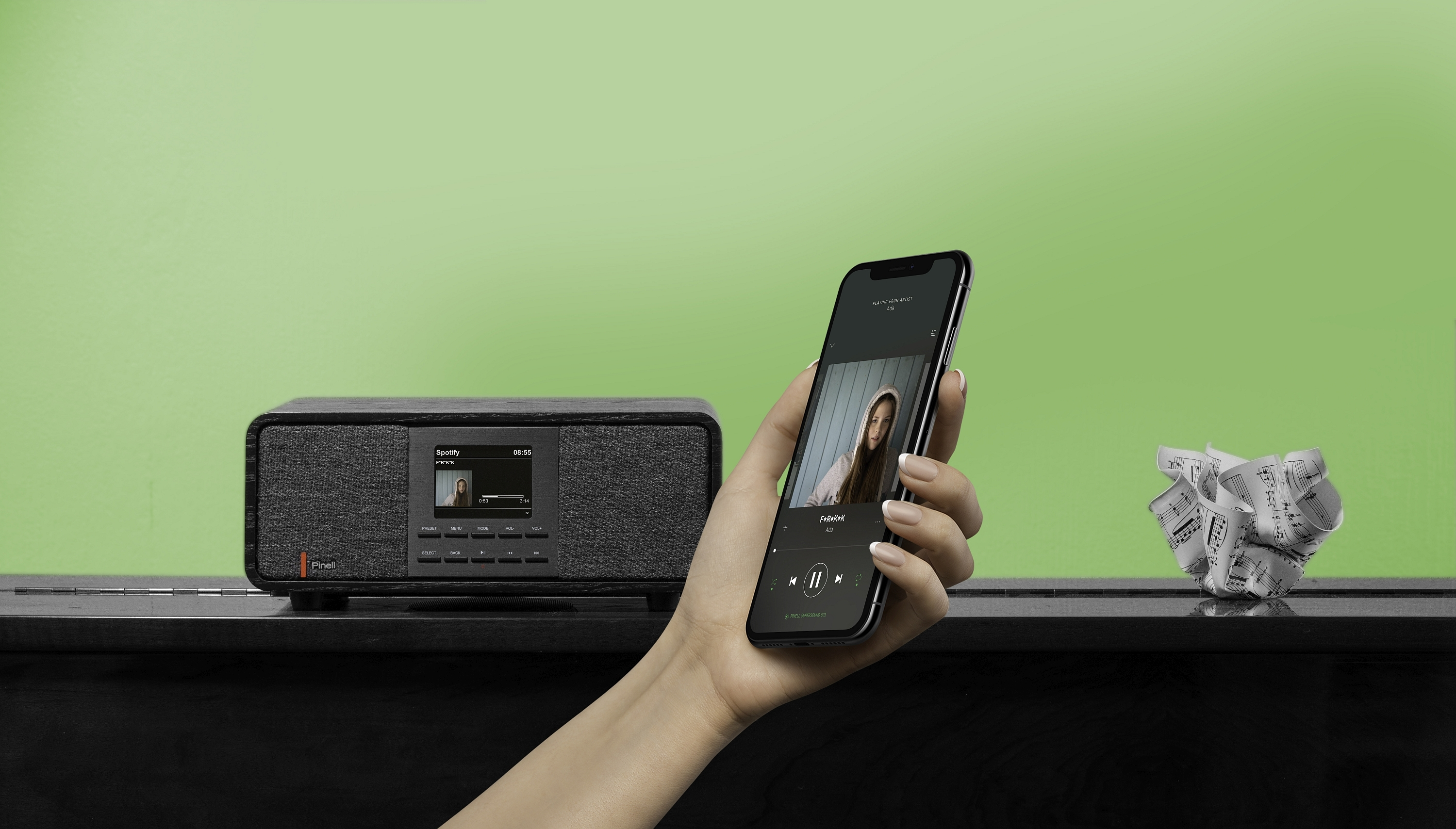 A hand holds a smartphone displaying a Spotify interface. A speaker and sheet music are on a black piano.