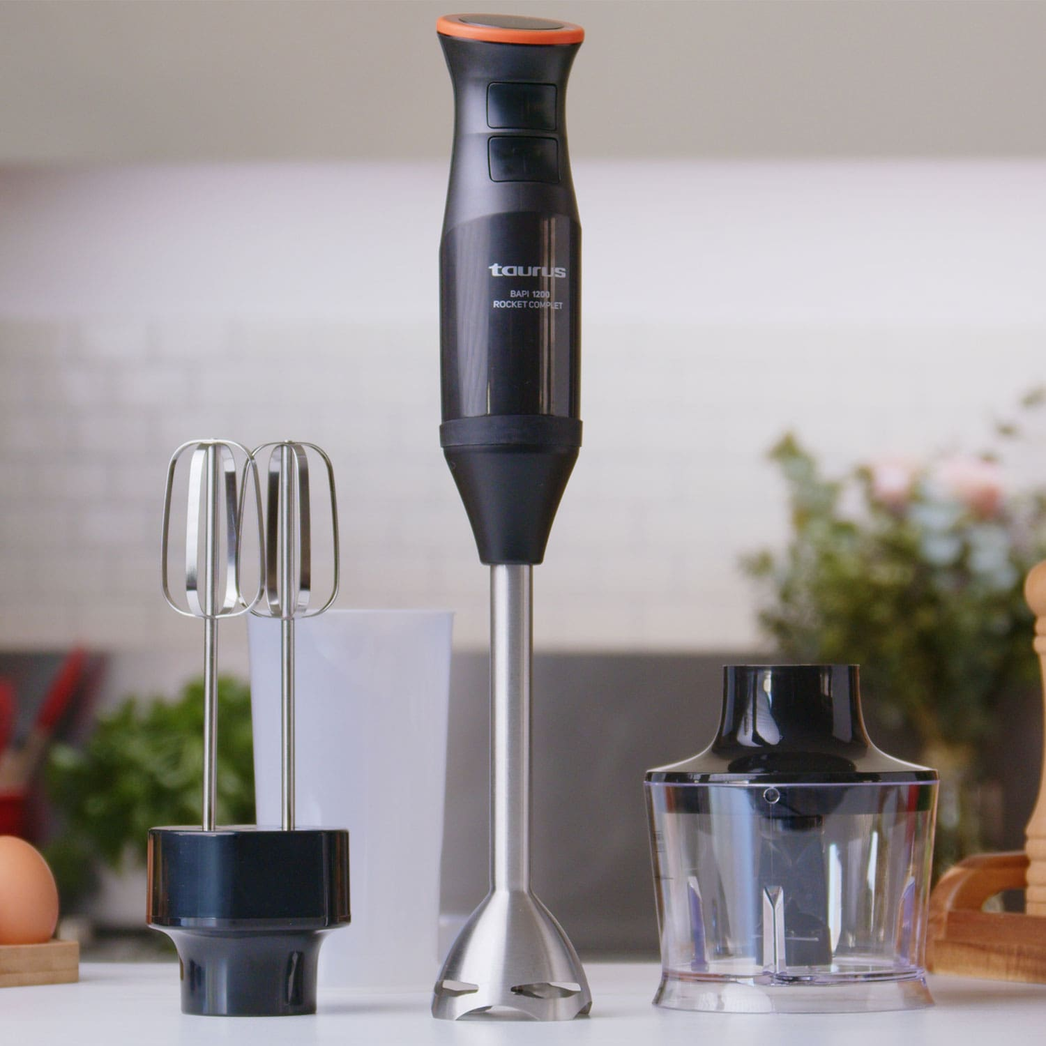 Una batidora de mano eléctrica, con accesorios, y accesorios sobre una superficie blanca frente a una cocina.
