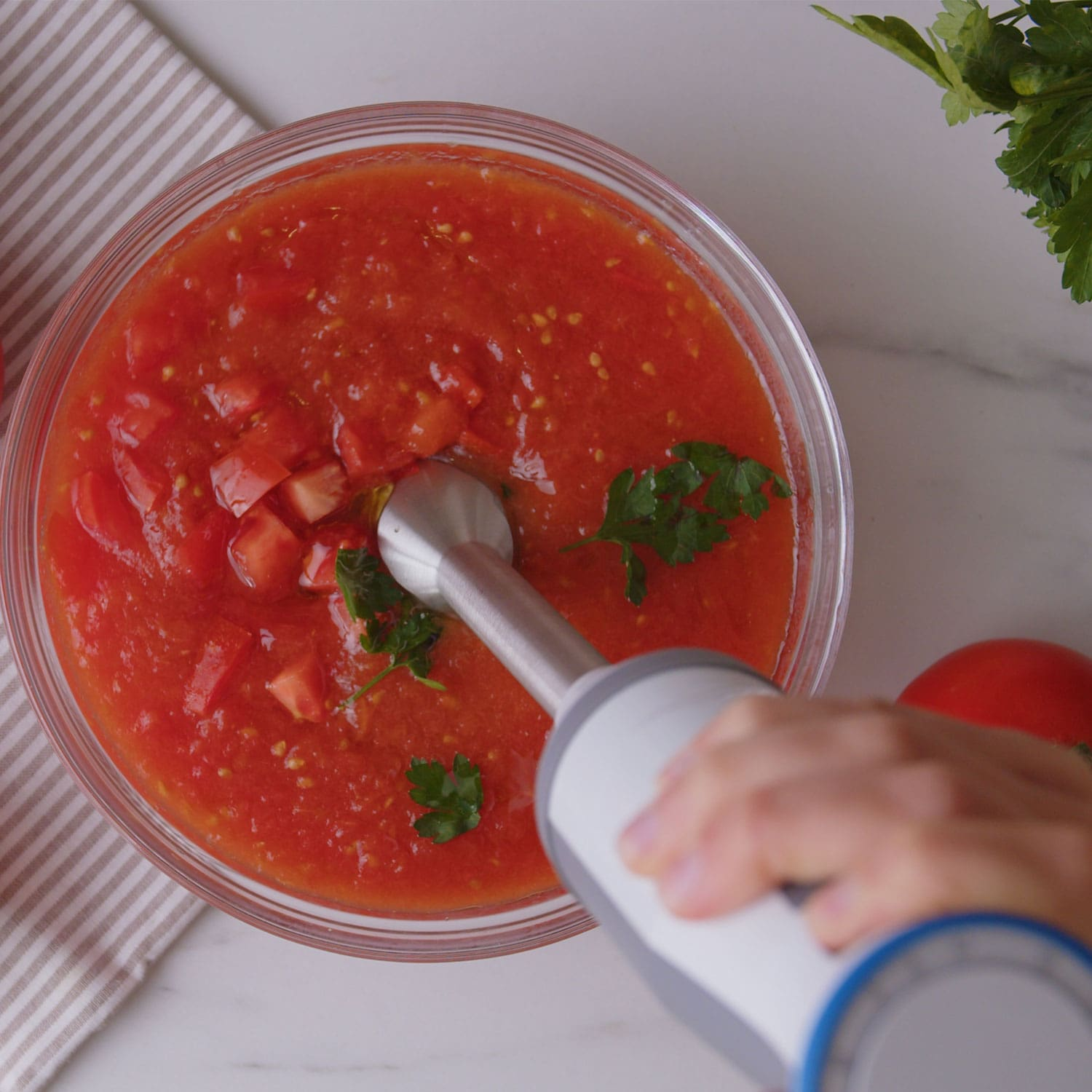 Mano mezclando sopa de tomate con una batidora de inmersión en un bol de cristal. Primer plano.