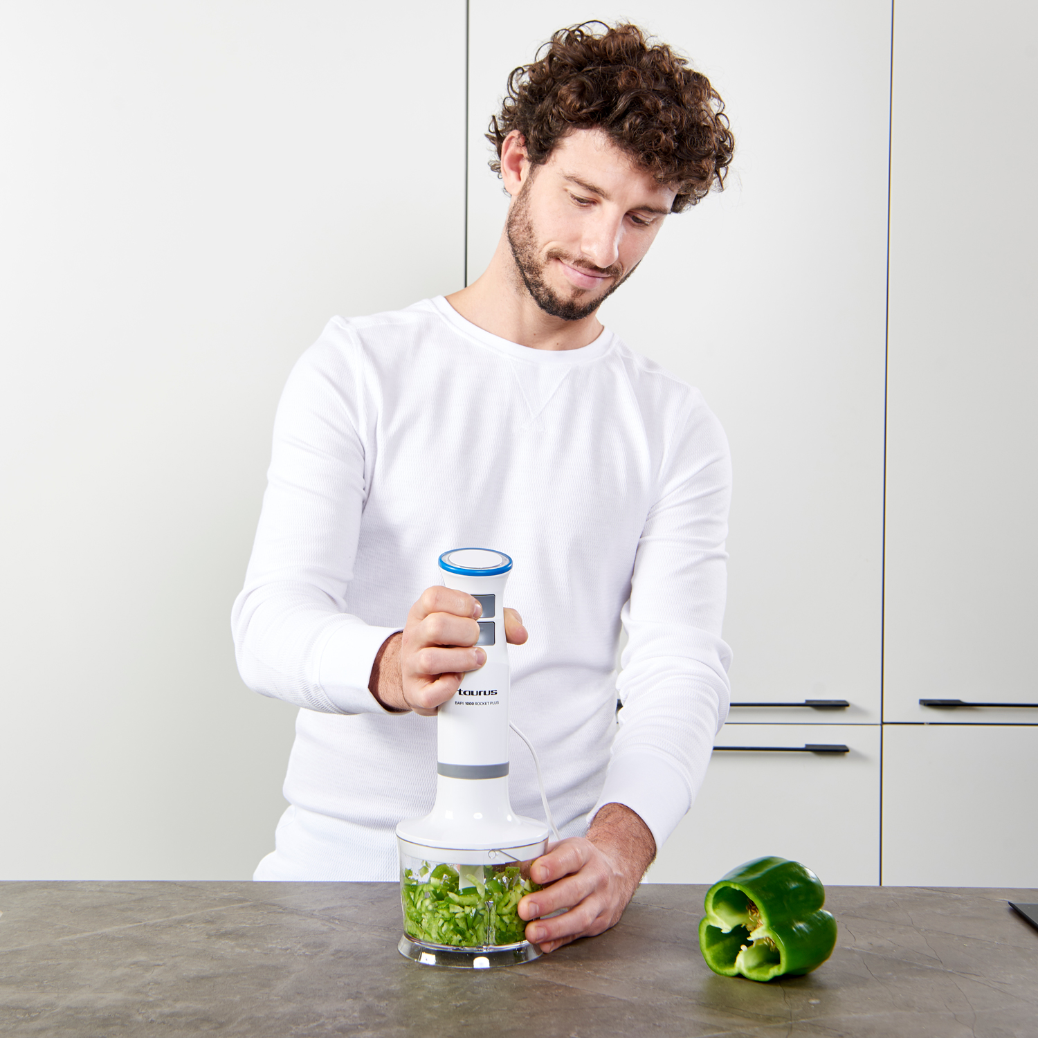 Hombre usando una batidora de mano con pimiento verde en un bol. Ambiente de cocina con armarios blancos.