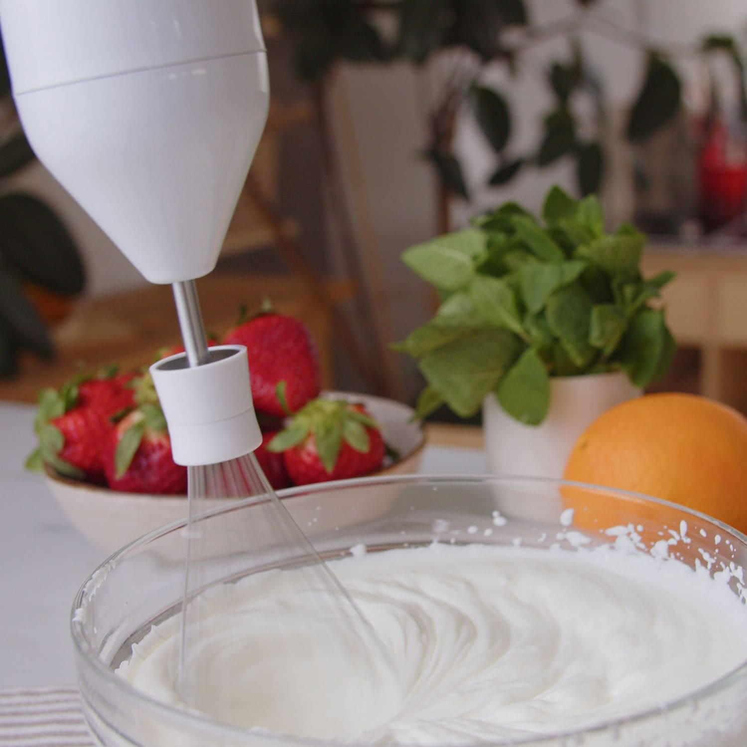 Una batidora de mano blanca mezclando crema en un bol de vidrio, fresas y una naranja al fondo.