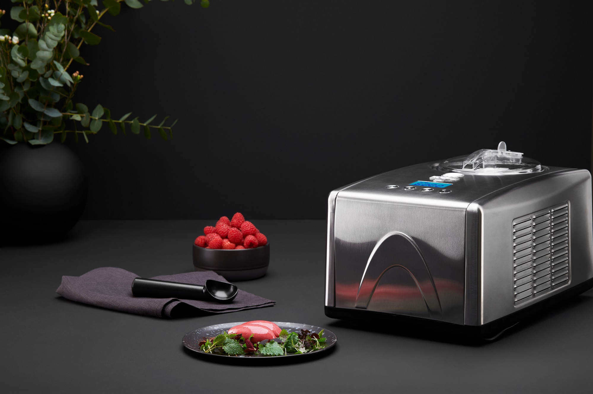 Silberner Eisbereiter mit schwarzem Löffel, Himbeeren in einer Schüssel und Dessertteller mit dunklem Hintergrund.