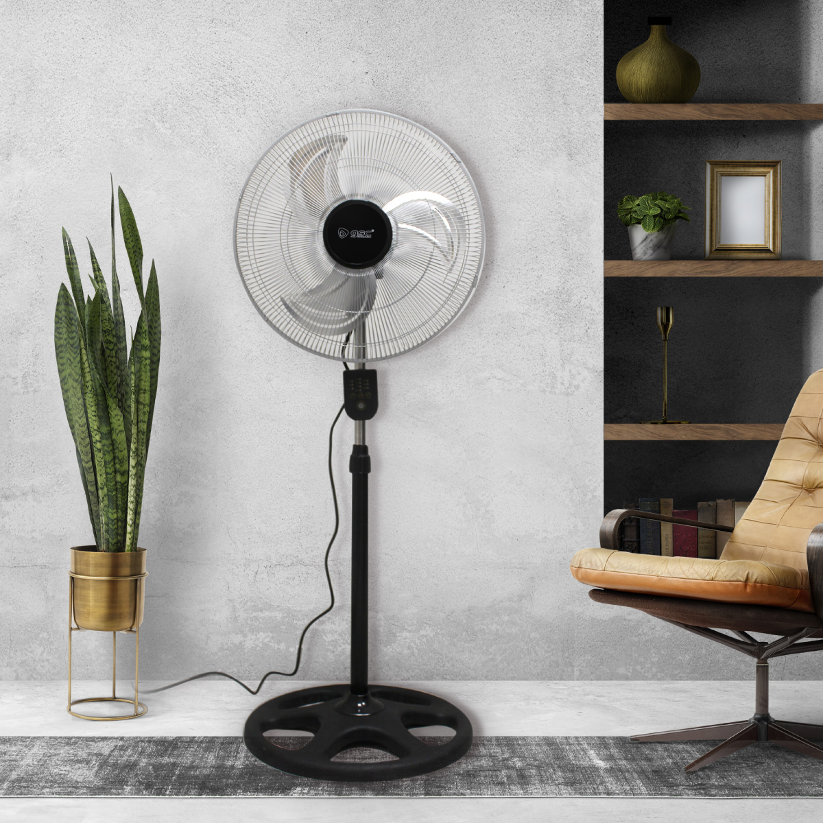 Un ventilador de pie negro y plateado está en una habitación. A la izquierda hay una planta y a la derecha una silla.
