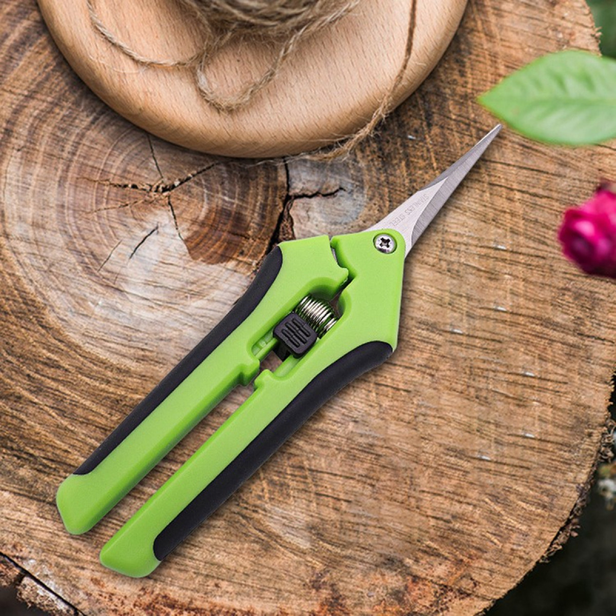 Grüne Gartenschere auf einem Holzscheit. Eine rosa Rose ist im Hintergrund sichtbar.