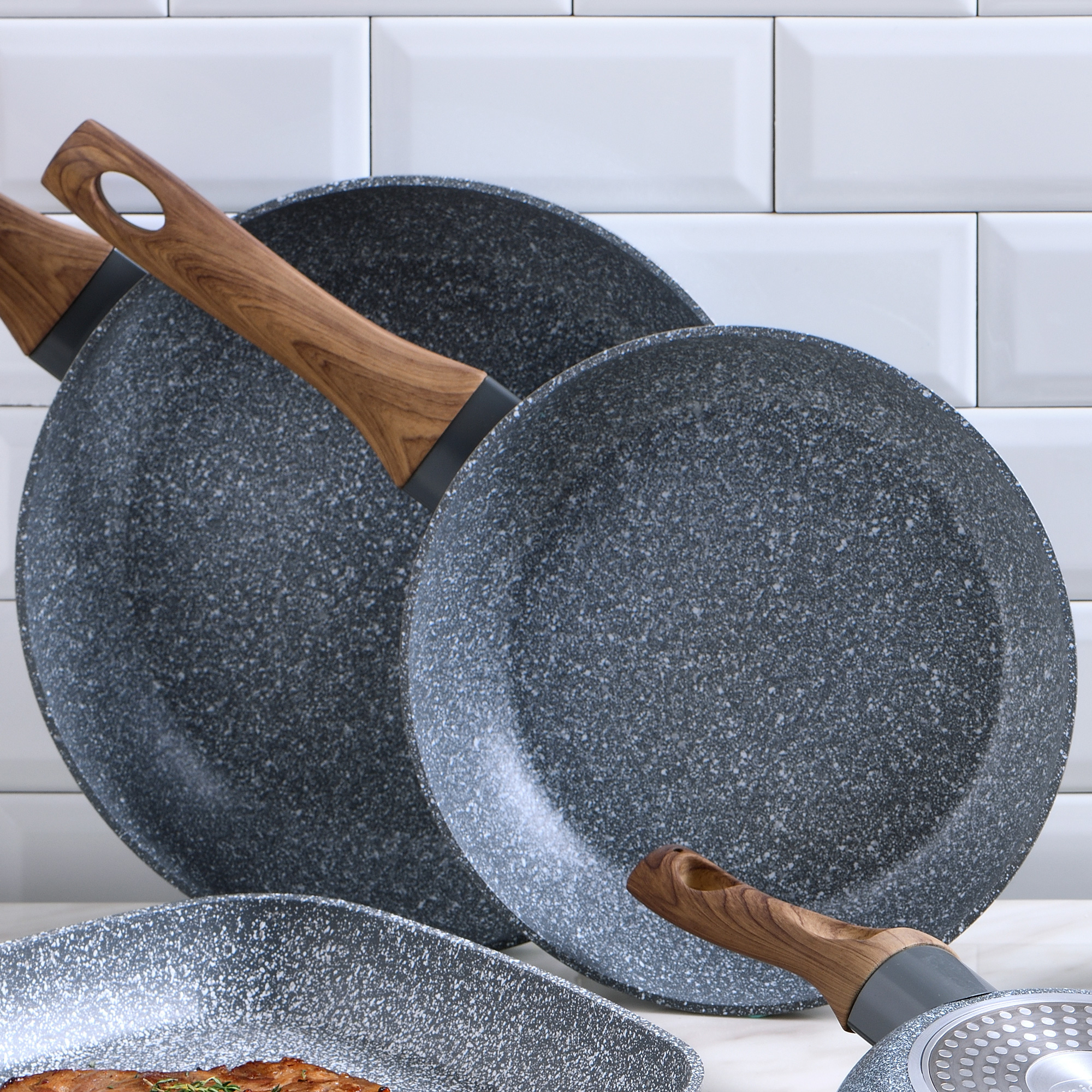 Tres sartenes con superficies moteadas grises. Mangos de madera. Fondo de azulejos blancos.