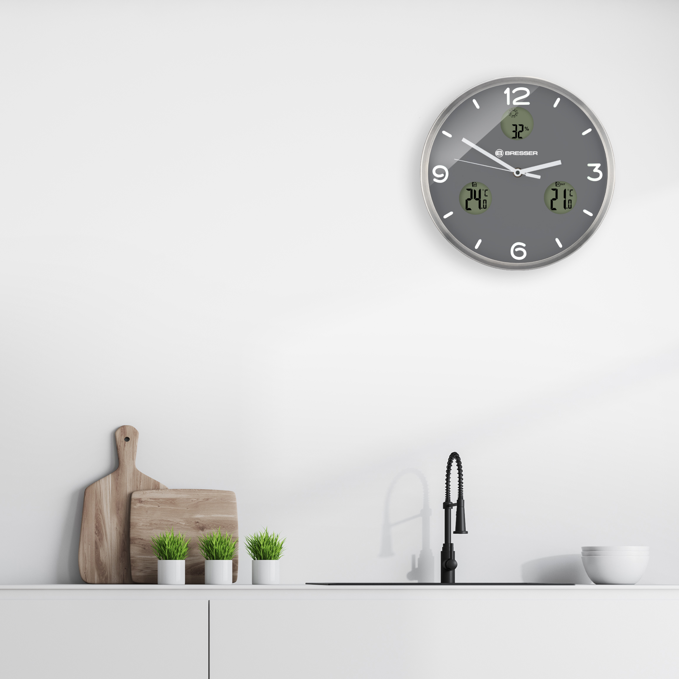 Reloj de pared gris con lecturas de temperatura y humedad. Ambiente de cocina con fregadero y grifo.