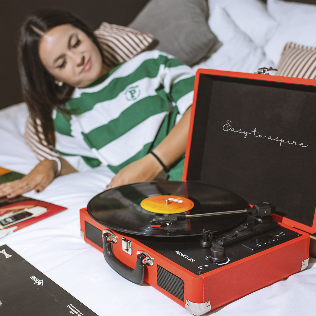 Kobieta leży na łóżku obok czerwonego gramofonu. Płyta winylowa na talerzu.