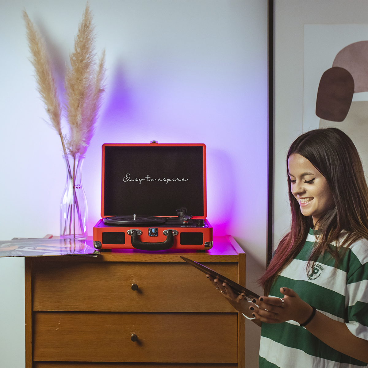 Kobieta uśmiecha się, trzymając płytę. Gramofon stoi na drewnianej szafce.